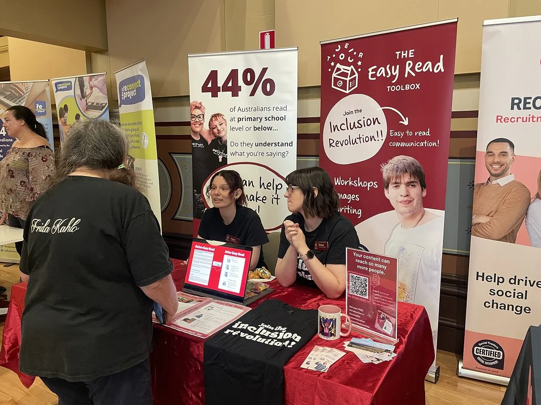 Two staff members from The Easy Read Toolbox sit at an expo stall speaking with a visitor, surrounded by banners promoting accessible communication and the Inclusion Revolution.