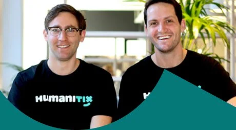 Two men in black Humanitix t-shirts smiling together outdoors.
