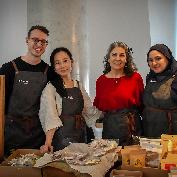 Four FoodLab Sydney team members smiling together behind a market stall table displaying packaged food products.