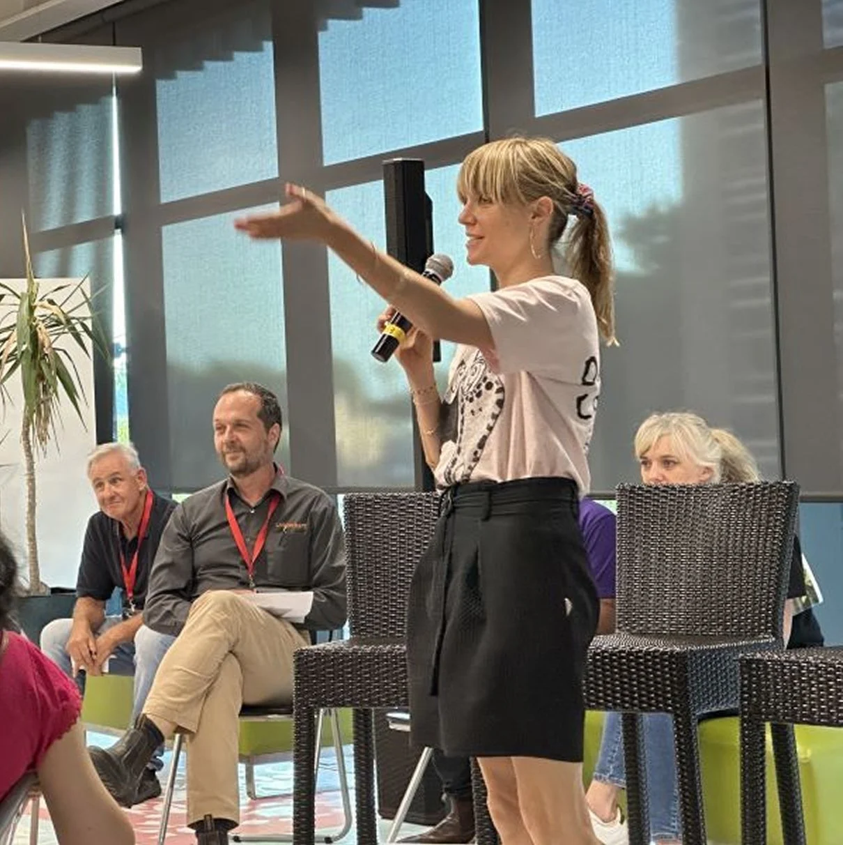 A woman holding a microphone addresses a seated panel at an indoor SECNA event, gesturing toward the audience.