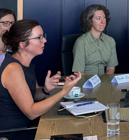 Two women seated at a boardroom table in conversation, with notepads and name placards in front of them.