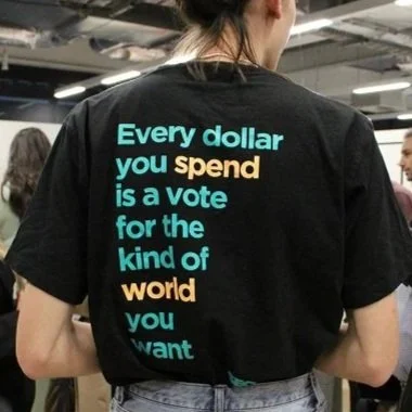 The back of a woman wearing a black SECNA shirt that reads "Every dollar you spend is a vote for the kind of world you want"