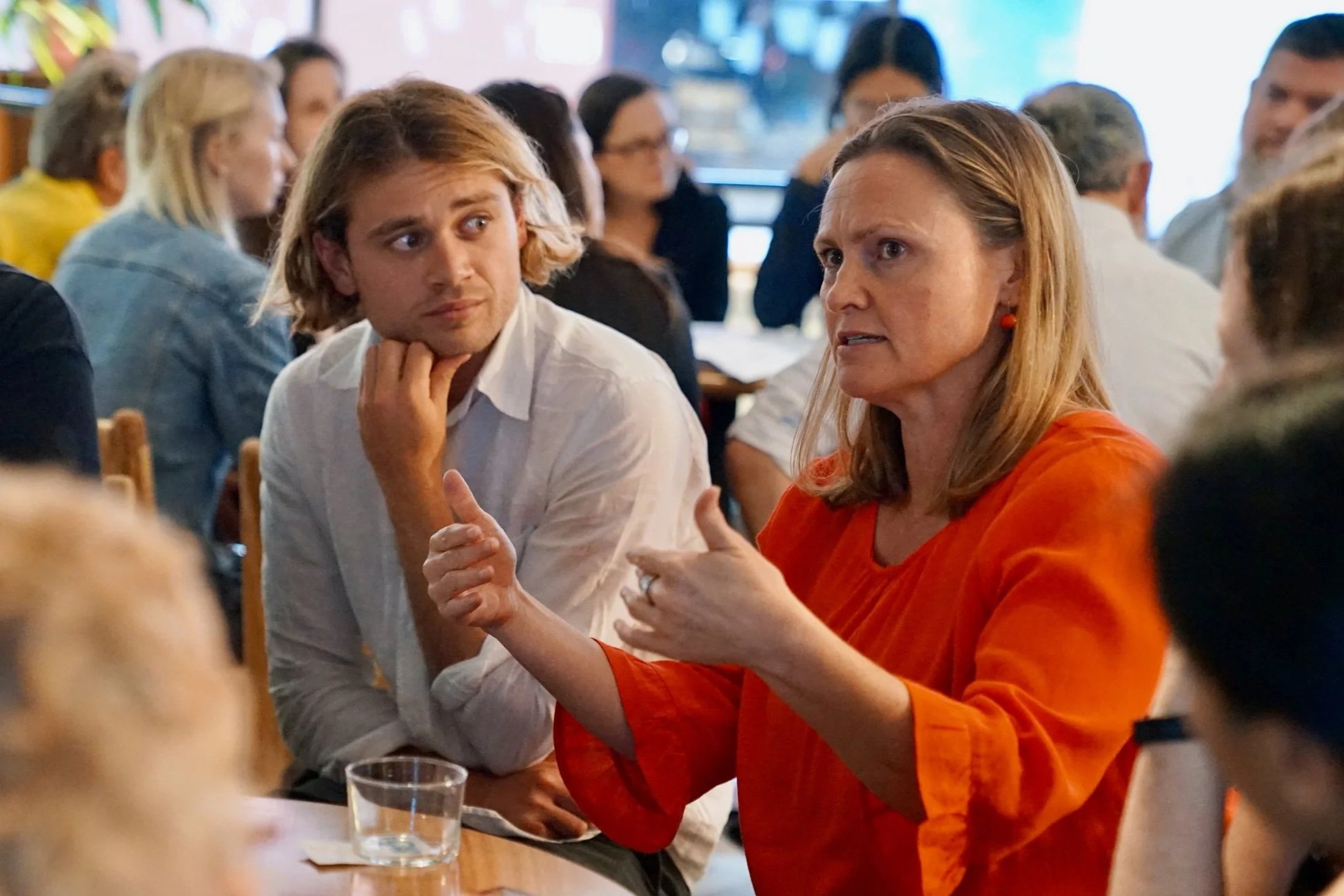 A woman in an orange top speaks expressively with hand gestures at a table, while a young man beside her listens attentively, with other attendees visible in the background.