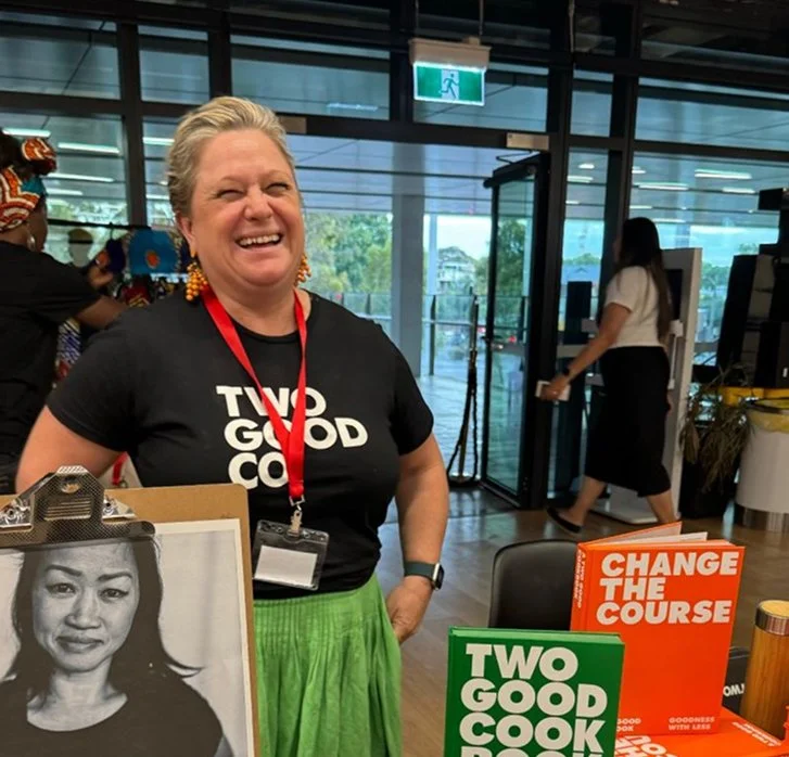 A smiling woman wearing a black 'Two Good Co' t-shirt and green skirt, holding materials at what appears to be a community or charity event, with 'Two Good Co' branded books visible in the foreground.