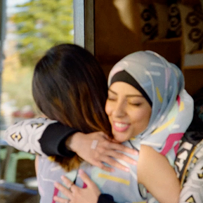 Two women sharing a warm embrace, one wearing a hijab, both smiling.