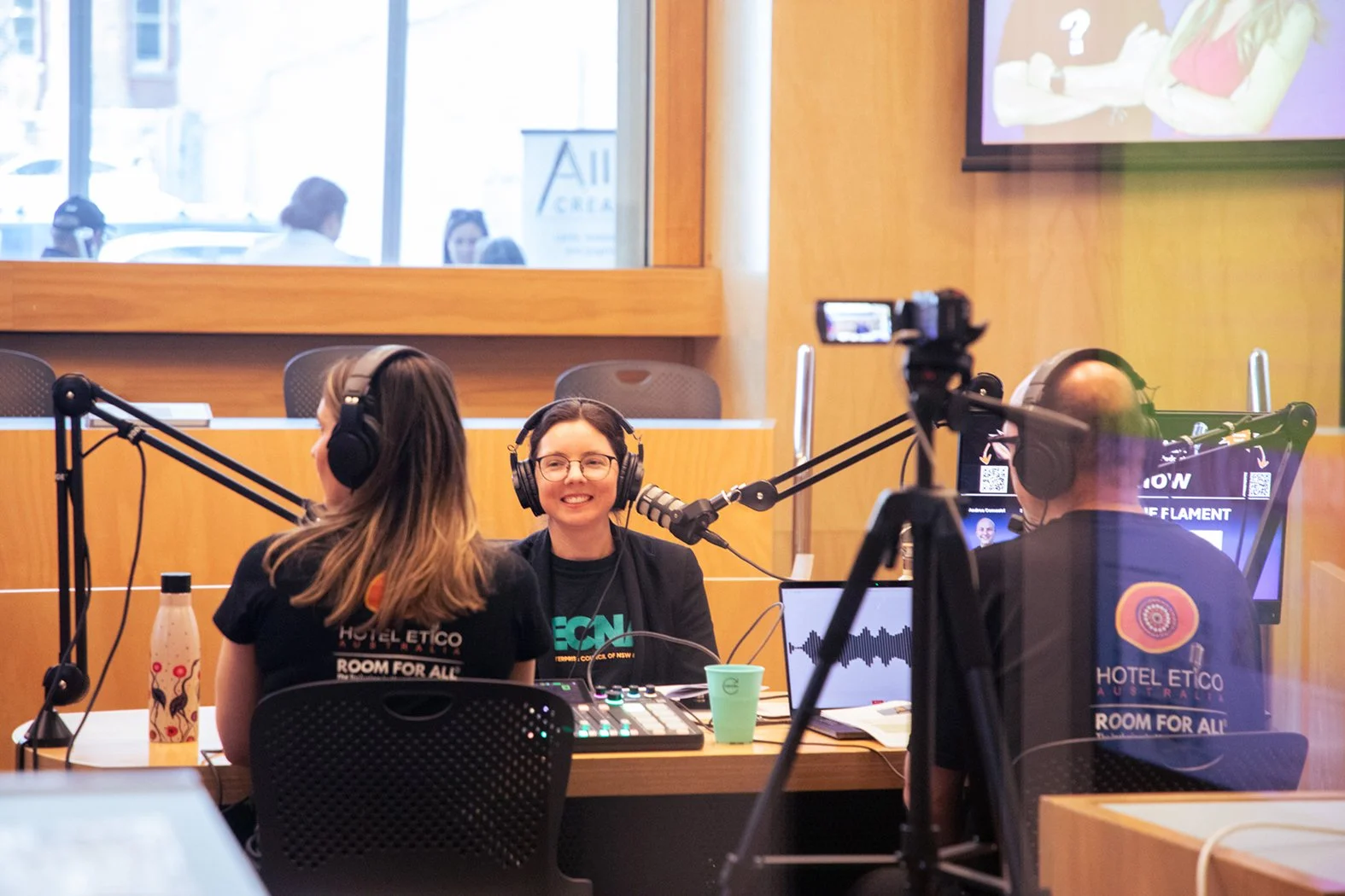 Two people wearing headphones and shirts that read "Hotel Etico: Room for all" on the back, record a podcast at a desk setup with microphones, a mixing board, and a camera, with Kylie (CEO of SECNA) smiling toward the other.