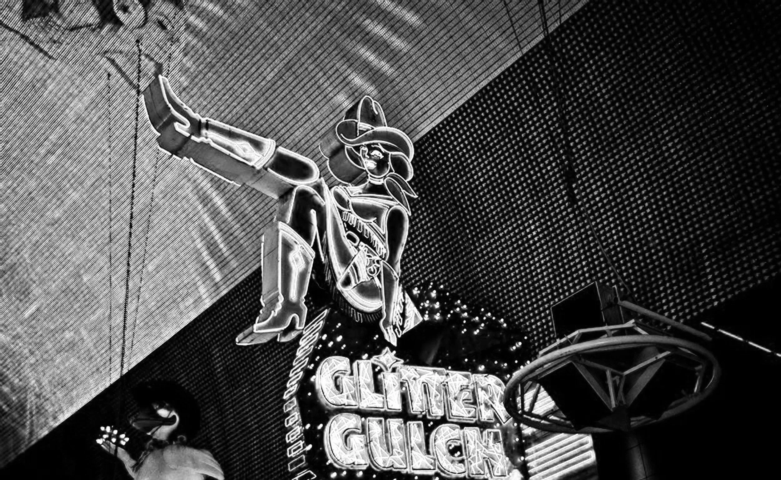 Neon sign of a cowboy figure above a venue entrance, with parts of a sign reading 'GUITAR' visible, against a textured wall and arched ceiling.