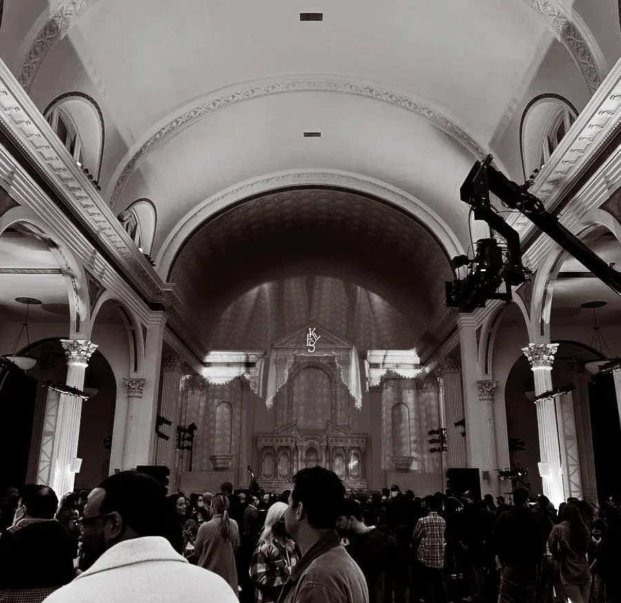 Black and white photo of a large church or cathedral interior with a high arched ceiling, columns, and a crowd of people gathered inside, with lighting equipment overhead.