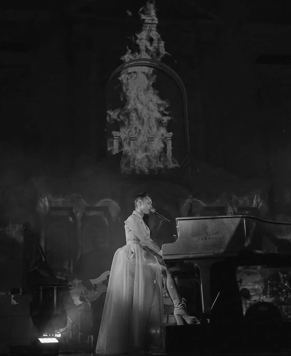 A woman in a long dress sitting at a Yamaha piano, singing into a microphone on a dimly lit stage with a projected image of fire and an ornate fountain in the background.