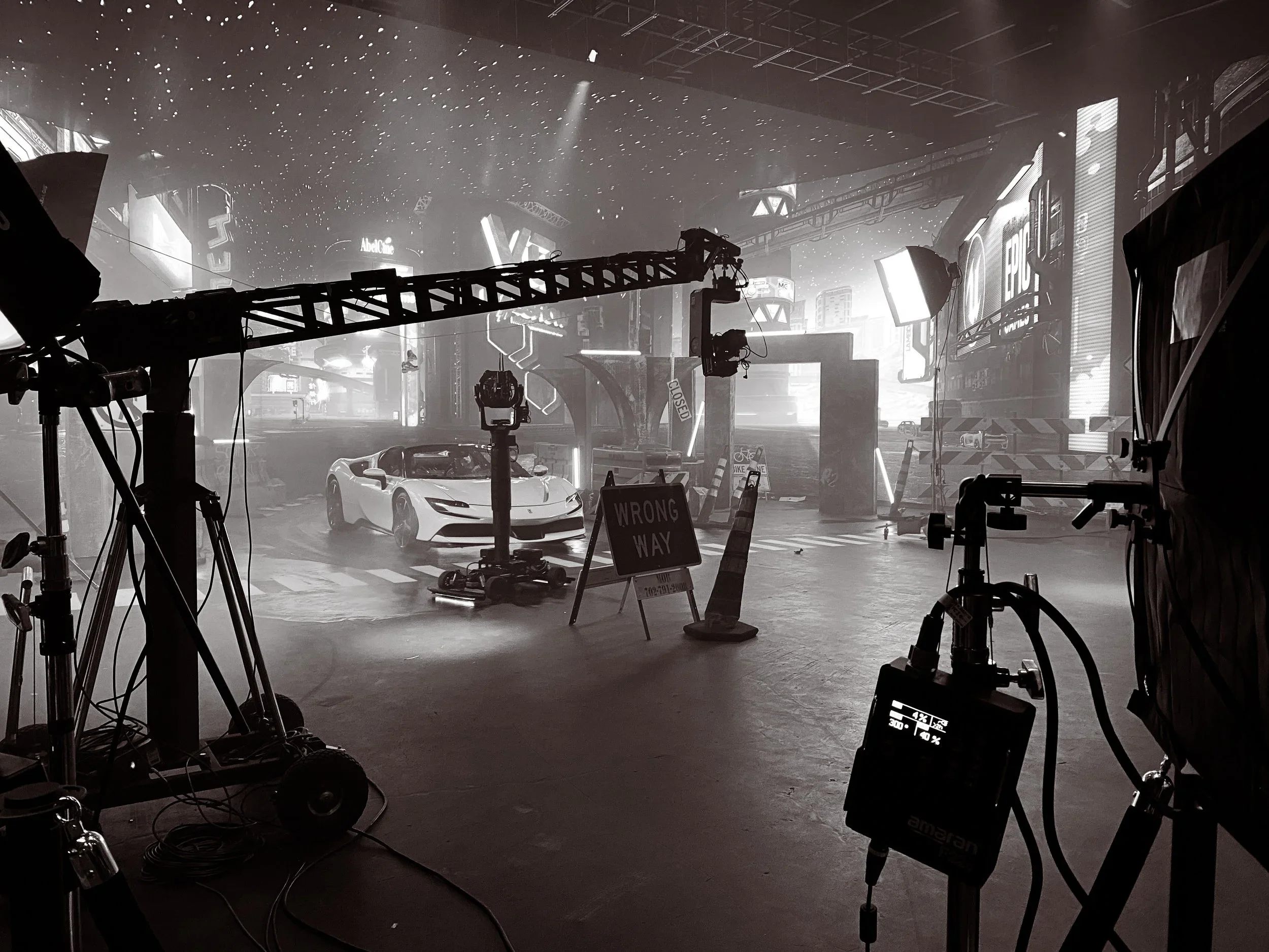 Film or photography set with cameras, lighting equipment, and a white sports car in a cityscape background with illuminated signs.