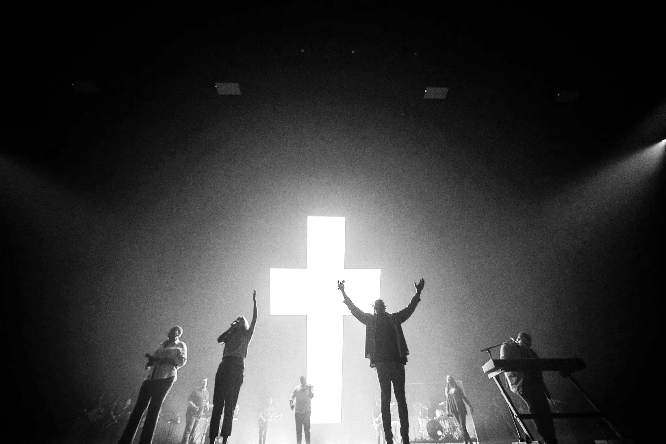 A black and white photo of a live music performance with a cross illuminated in the background. Several musicians are on stage, some with raised hands, including a lead singer with arms outstretched and others playing guitars and keyboards.