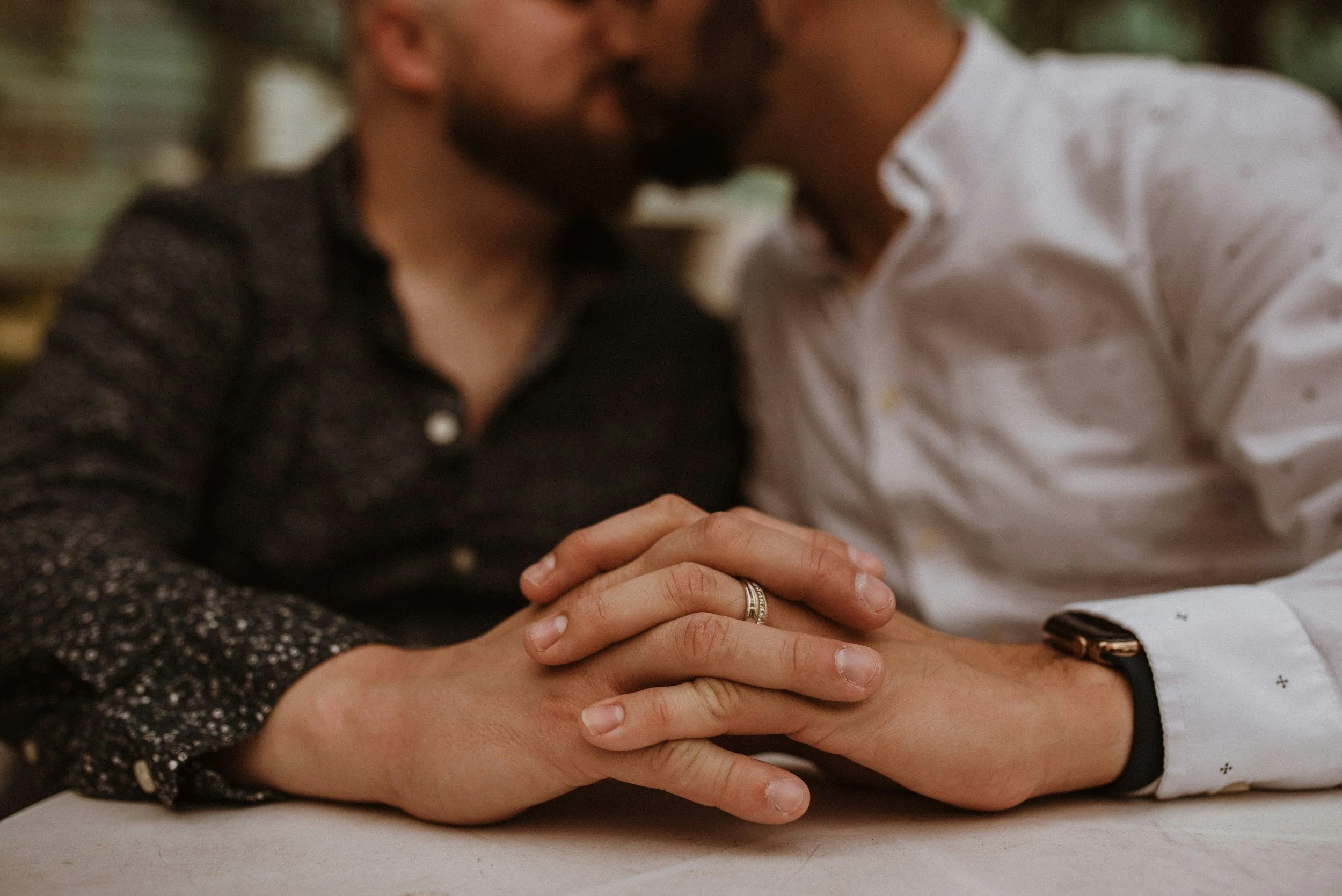 Two men are kissing and holding hands with wedding rings visible on their fingers.