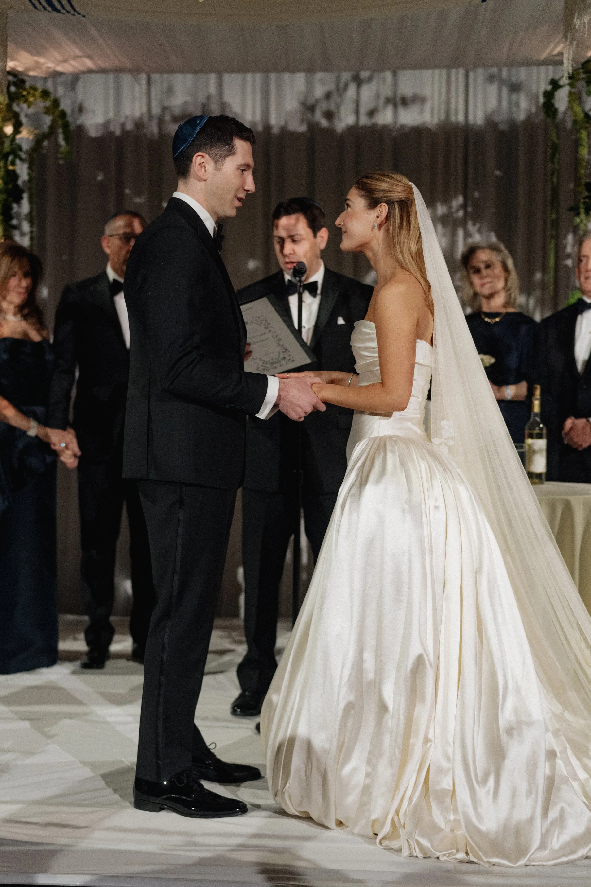 Bride and groom during their wedding ceremony, exchanging vows, with officiant and guests in the background.