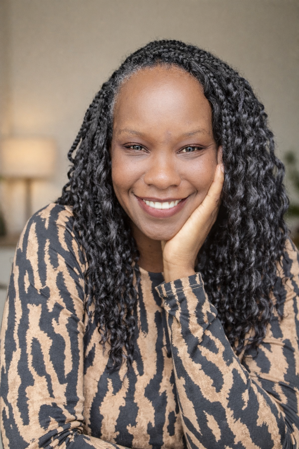 Close-up of a smiling African American woman with curly hair, resting her chin on her hand, wearing a black and beige animal print top, in a warmly lit indoor setting.
