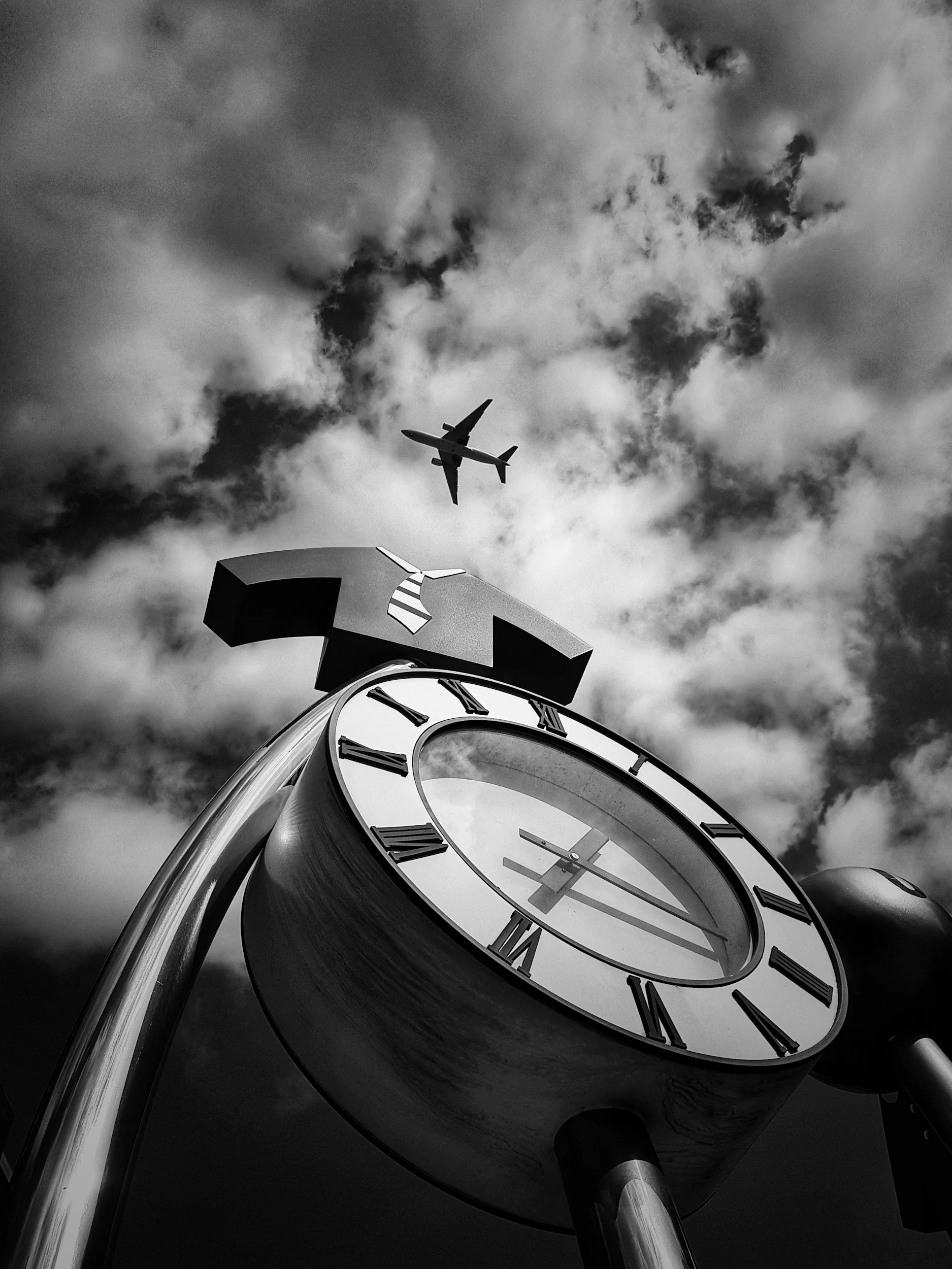 A black and white photo of a large clock with Roman numerals, viewed from below, with a sky filled with clouds and an airplane flying overhead.