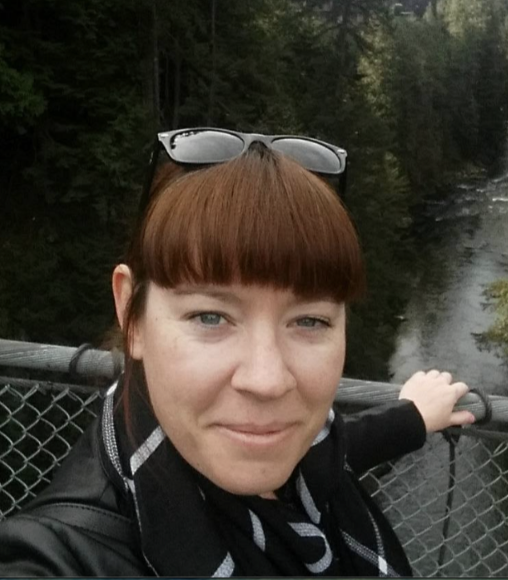Founder, Adrienne Marrone. She has auburn hair and sunglasses on her head. She's standing on a bridge with a metal railing, over a river surrounded by dense green trees.