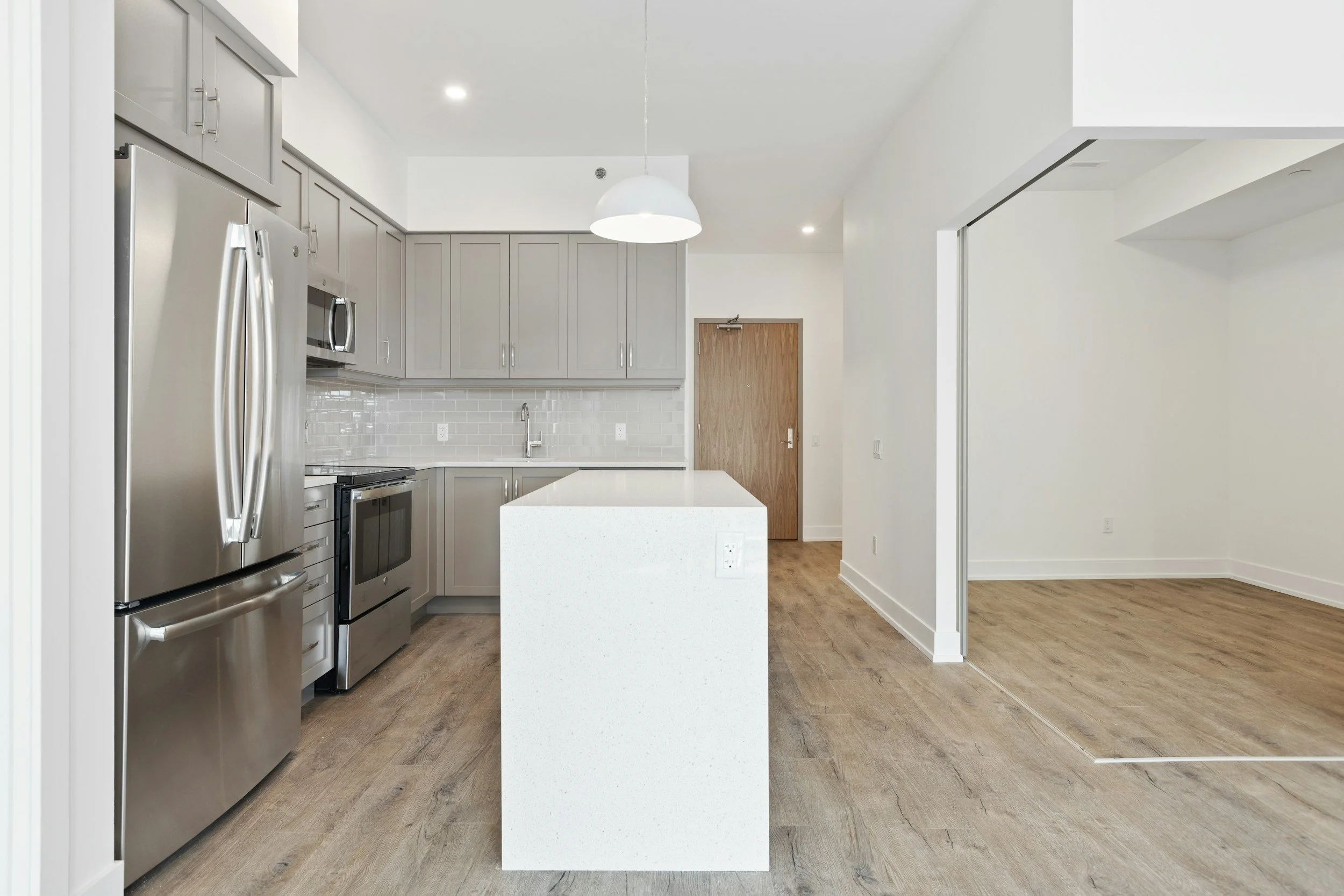 Modern kitchen with stainless steel appliances, gray cabinets, white island, wood flooring, and white walls.