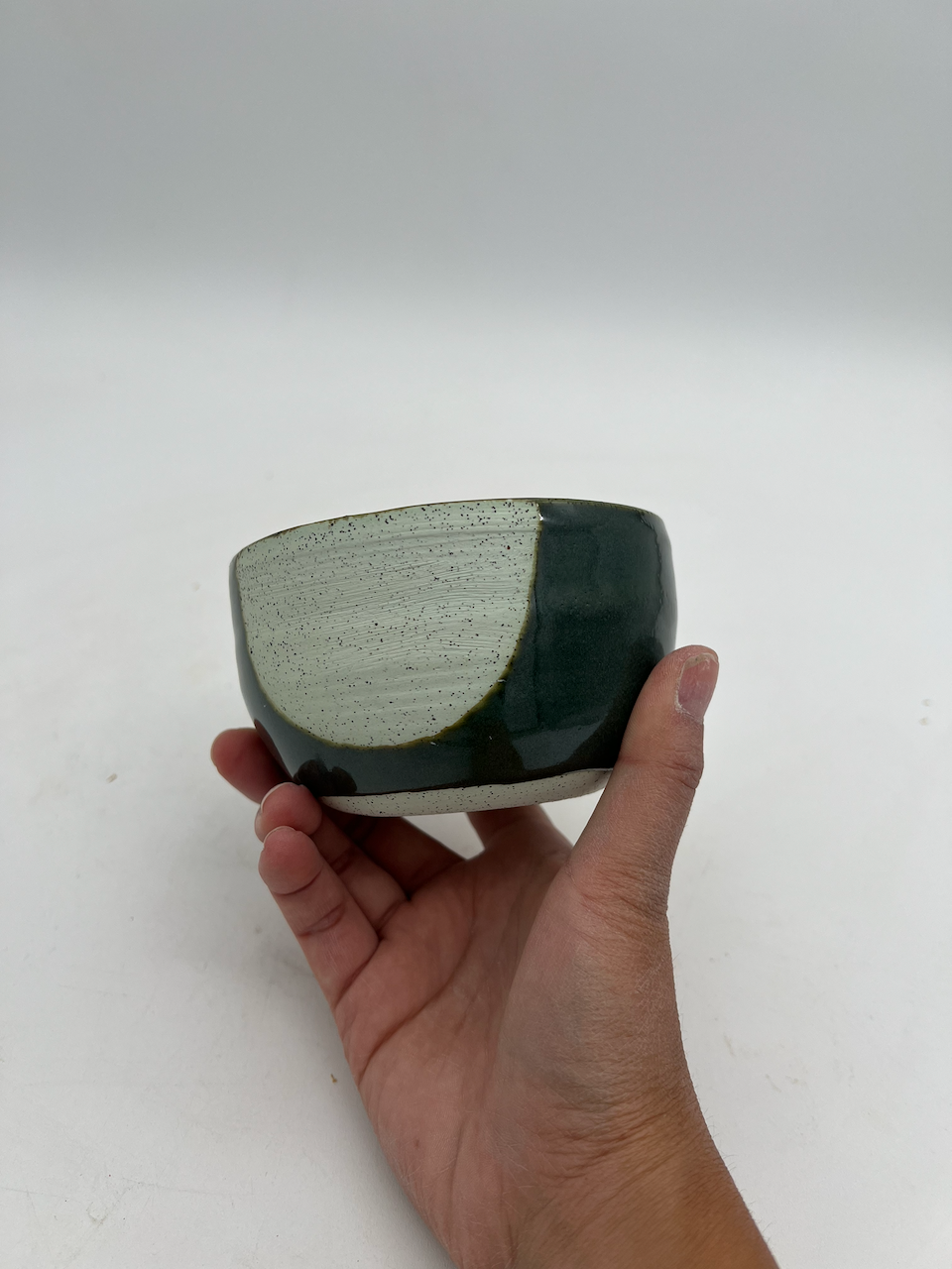 A ceramic bowl with a green glaze and plain clay, held in a person's hand against a plain white background.