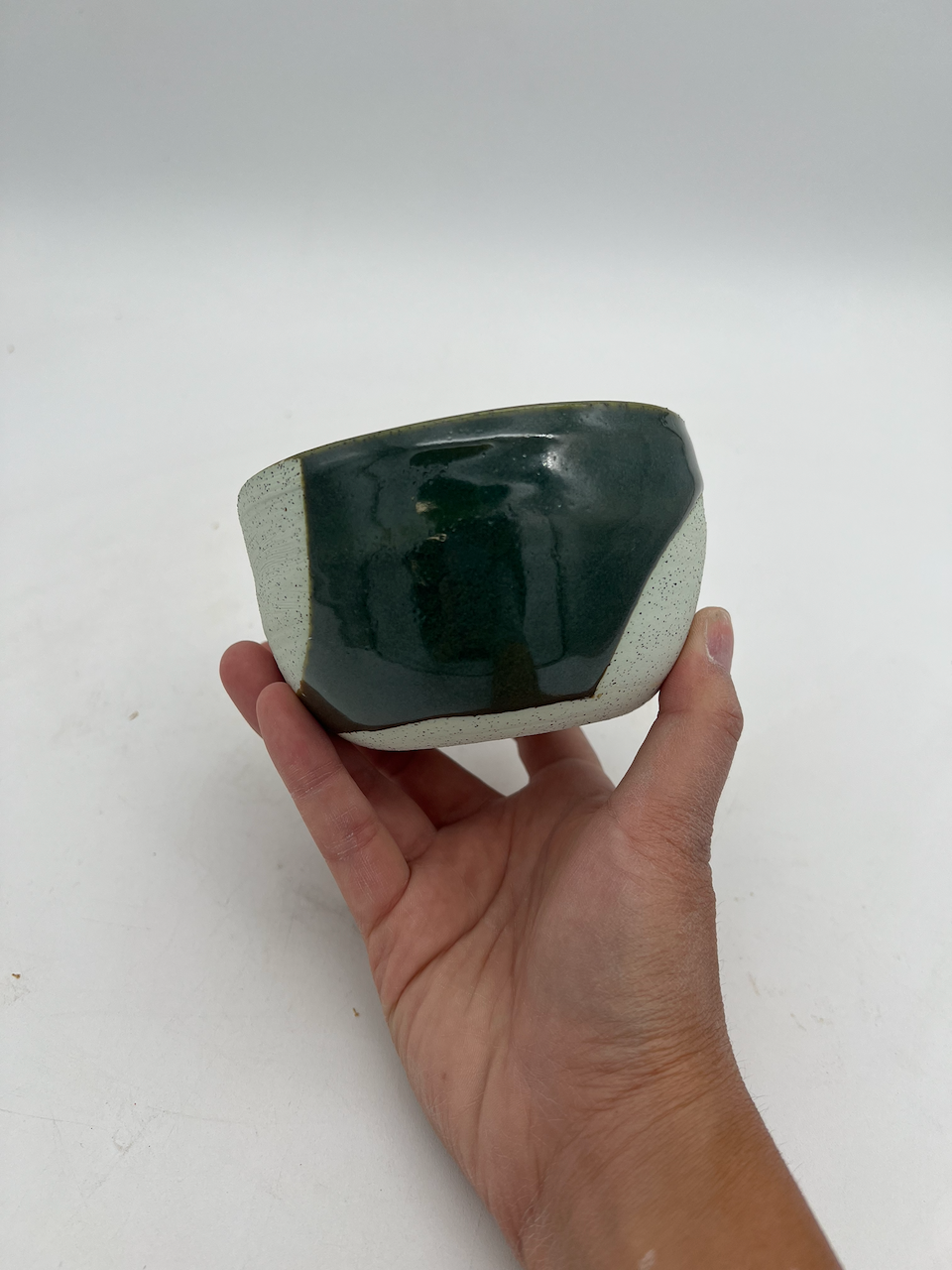 Hand holds a ceramic bowl with dark green and plain clay against a plain white background.