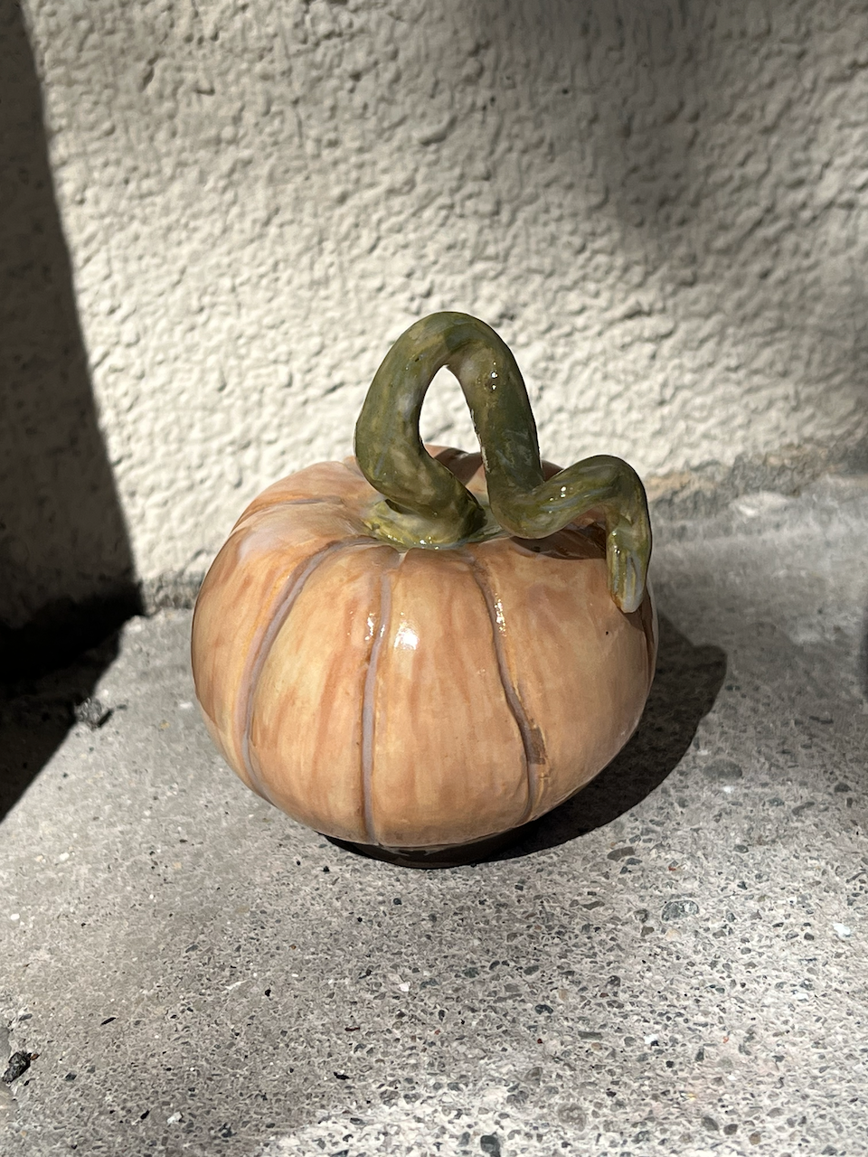 A pumpkin-shaped ceramic decorative object with a green vine handle on top, placed on a textured concrete surface outdoors.