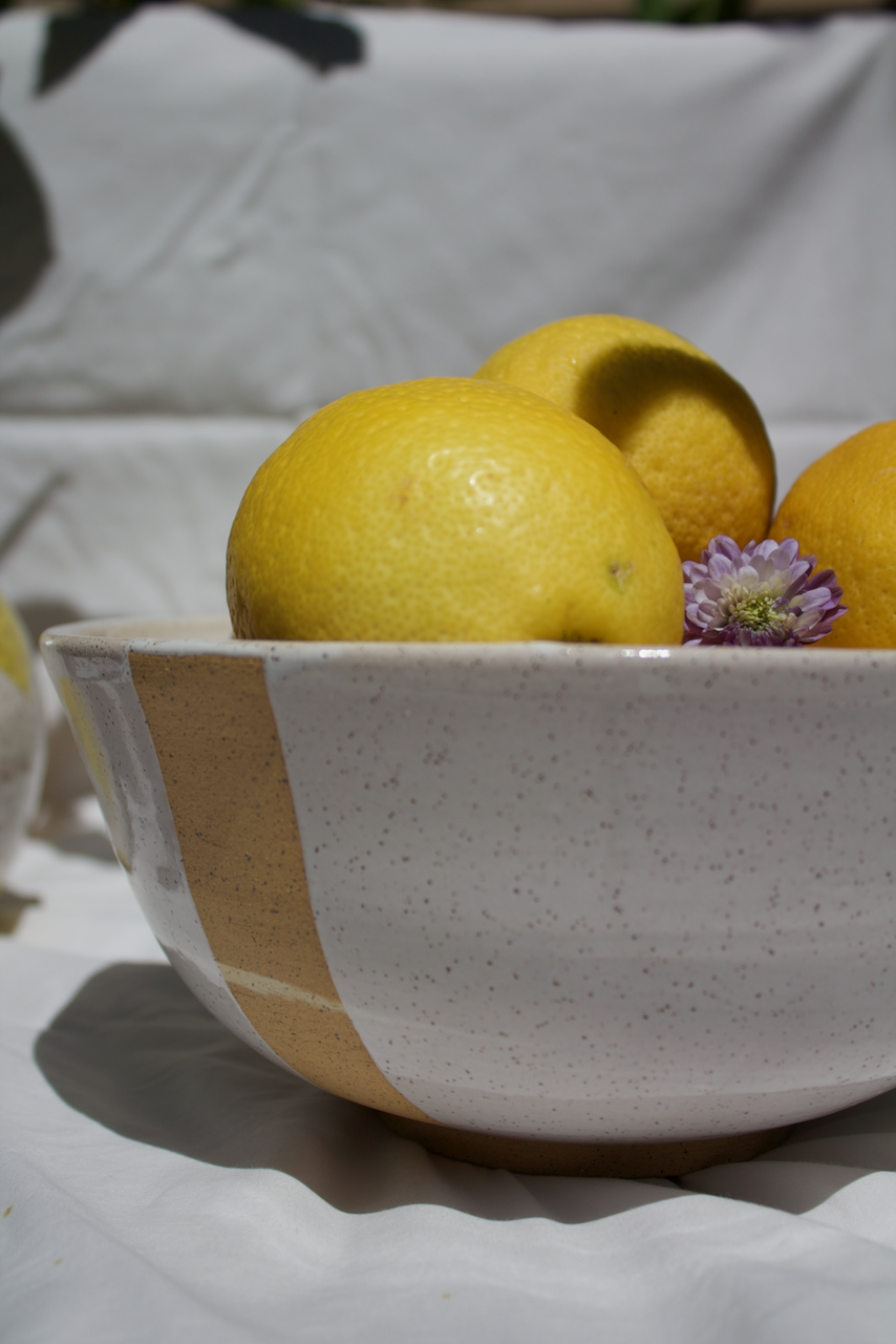 Close-up of a ceramic bowl filled with yellow lemons and a purple flower, set against a white cloth background.
