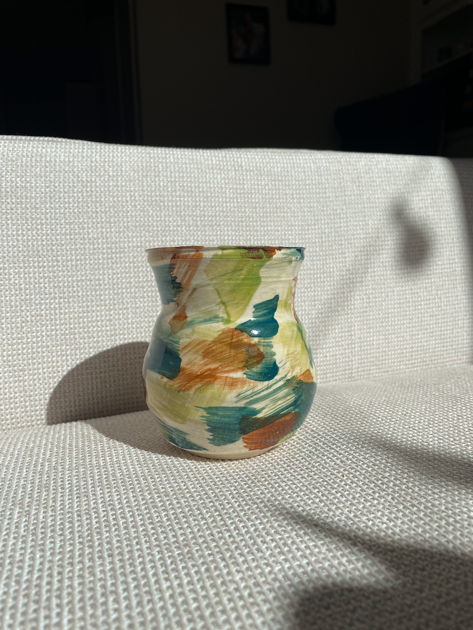 A small, rounded ceramic vase with an abstract, colorful brushstroke pattern in shades of green, blue, and brown, sitting on a textured white fabric surface in sunlight.
