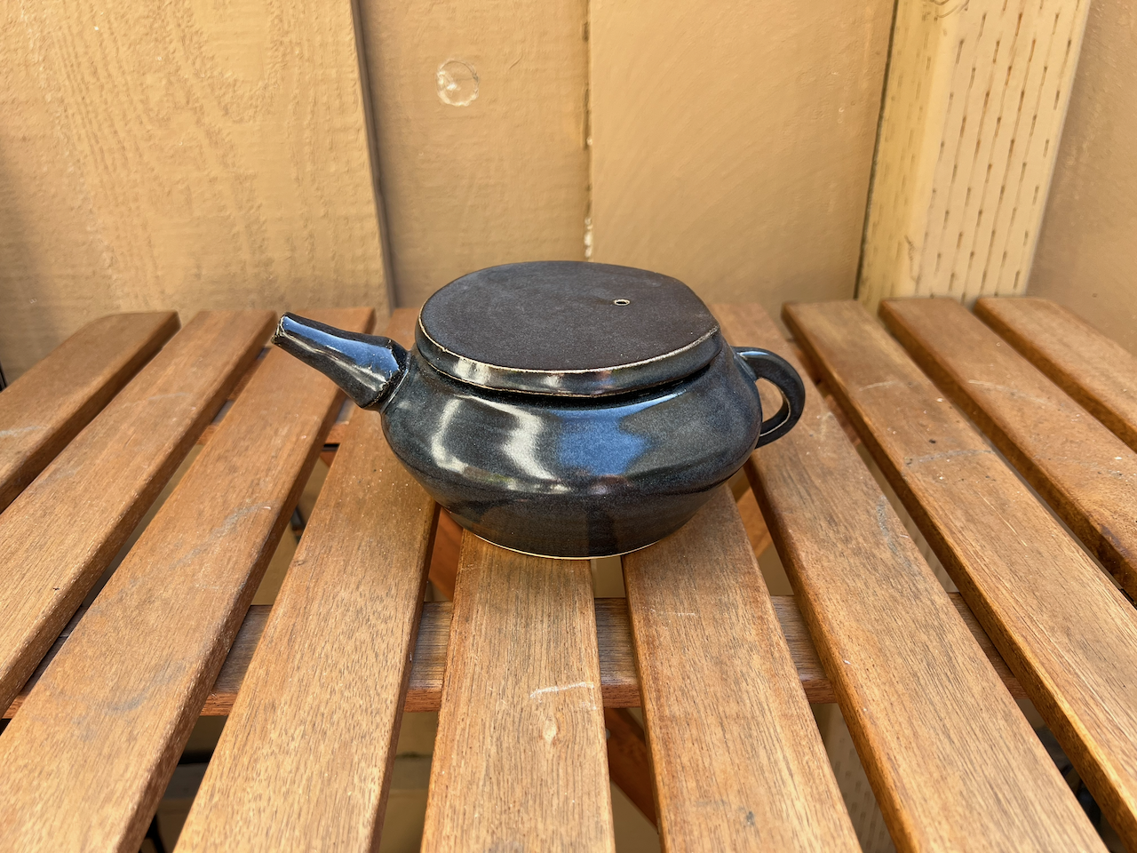 A black teapot with a round body, short spout, and handle, sitting on a wooden outdoor table.