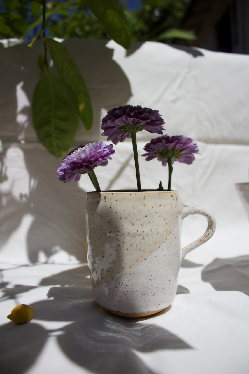 A ceramic mug with a speckled off-white glaze holding three purple flowers, set against a white background and greenery.
