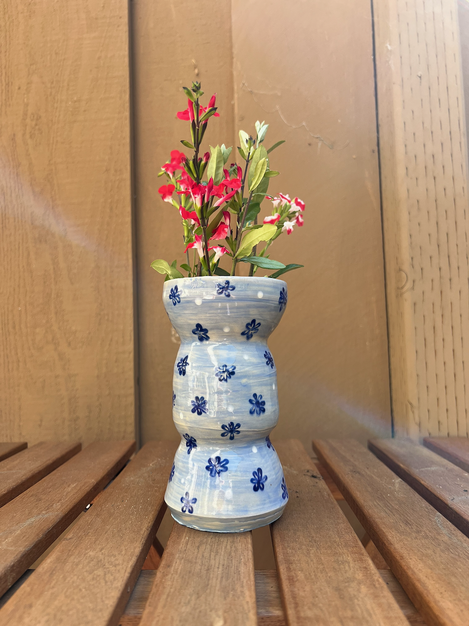 A white ceramic vase with blue flower patterns, containing pink and white flowers, placed on a wooden slatted surface against a wood-paneled background.
