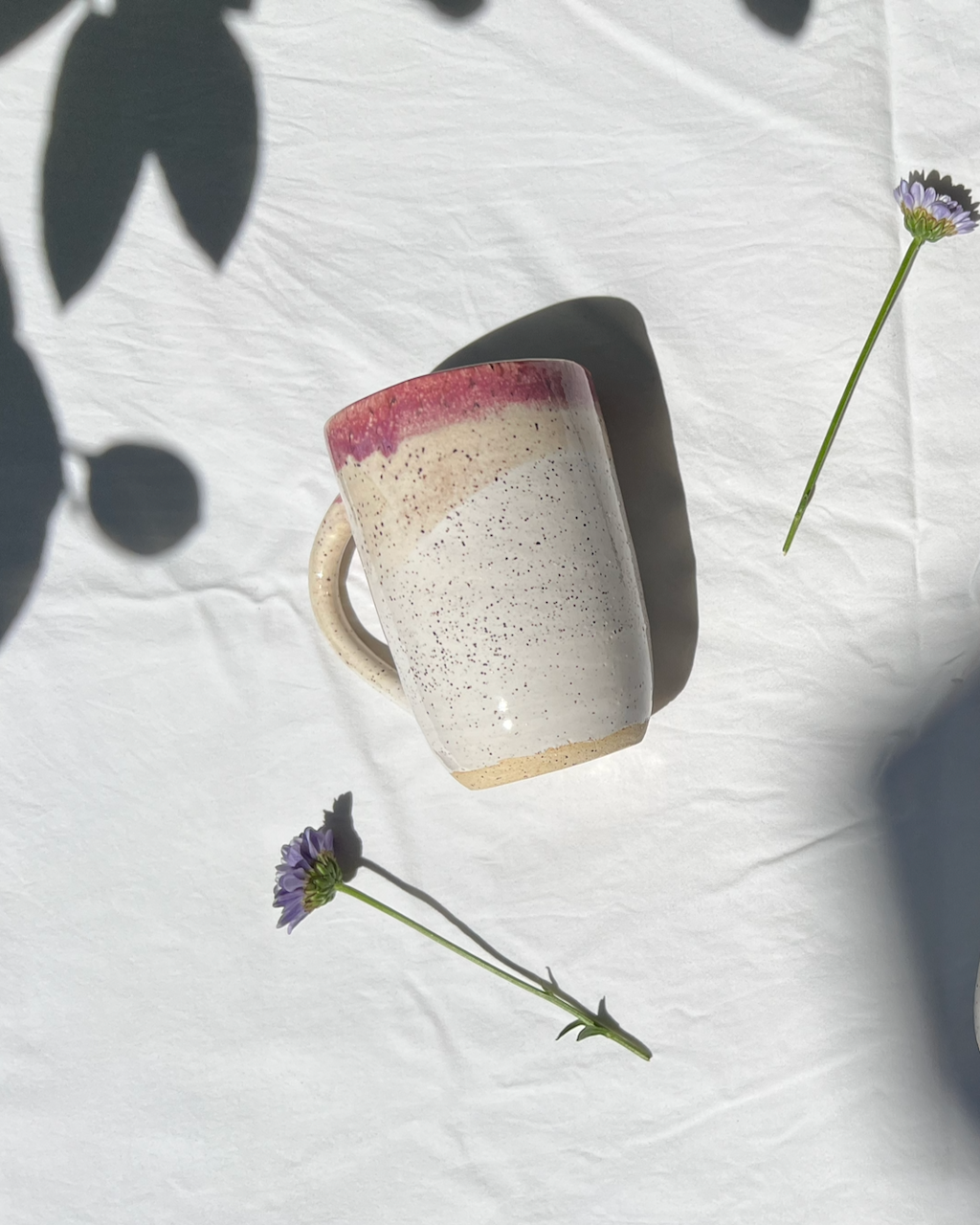A ceramic mug with a pink, beige, and cream gradient glaze, casting a shadow on a white textured surface. Two small purple flowers with green stems are nearby, with parts of leaves and another flower stem in the corner.