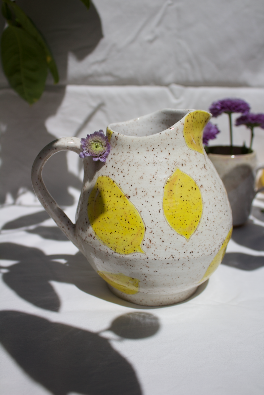 Ceramic pitcher with yellow lemon slice patterns, a small purple flower tucked on the handle, and reflected shadows, with purple flowers in background.