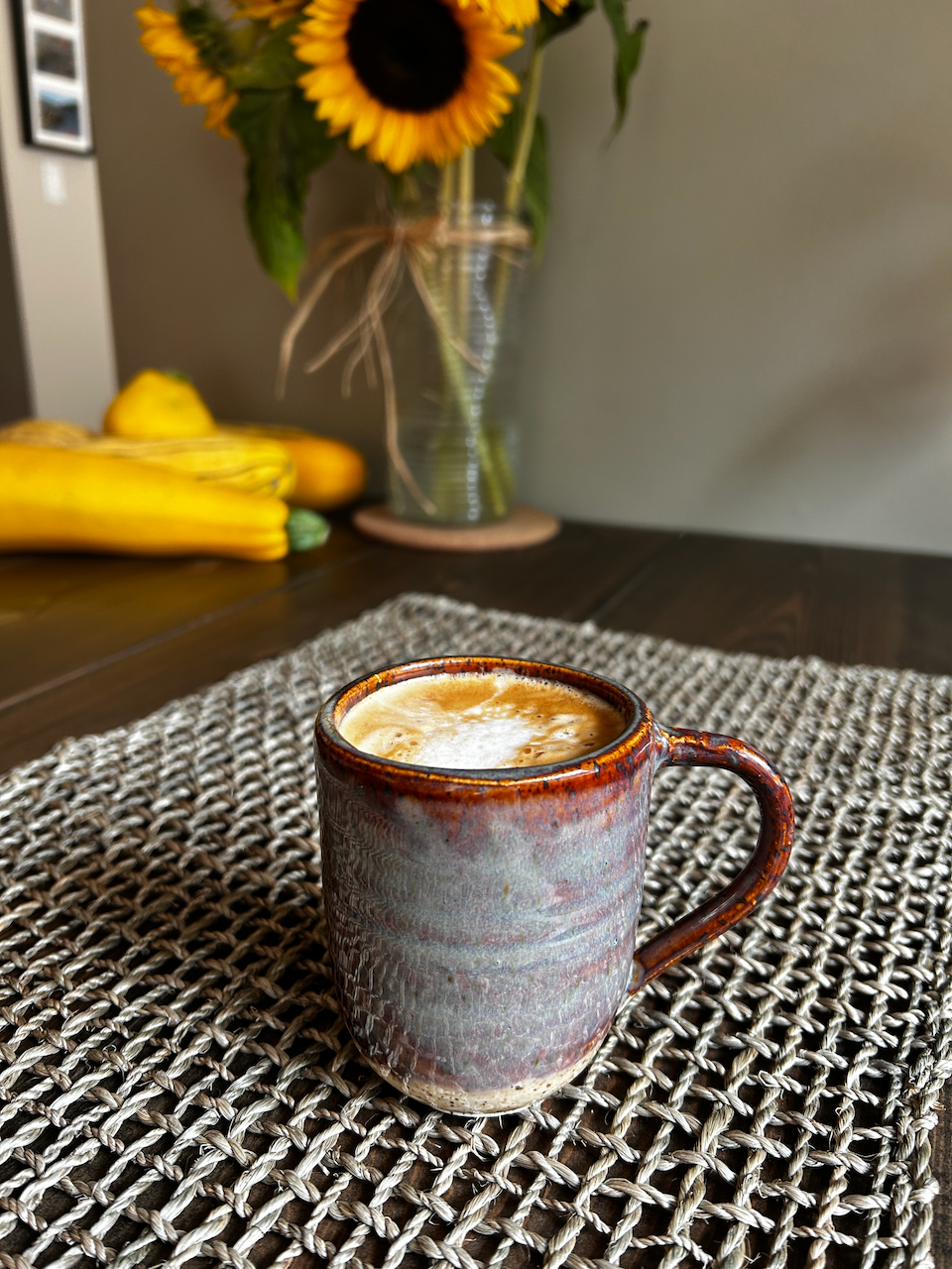 A ceramic mug filled with coffee on a woven placemat, with a vase of sunflowers in the background.