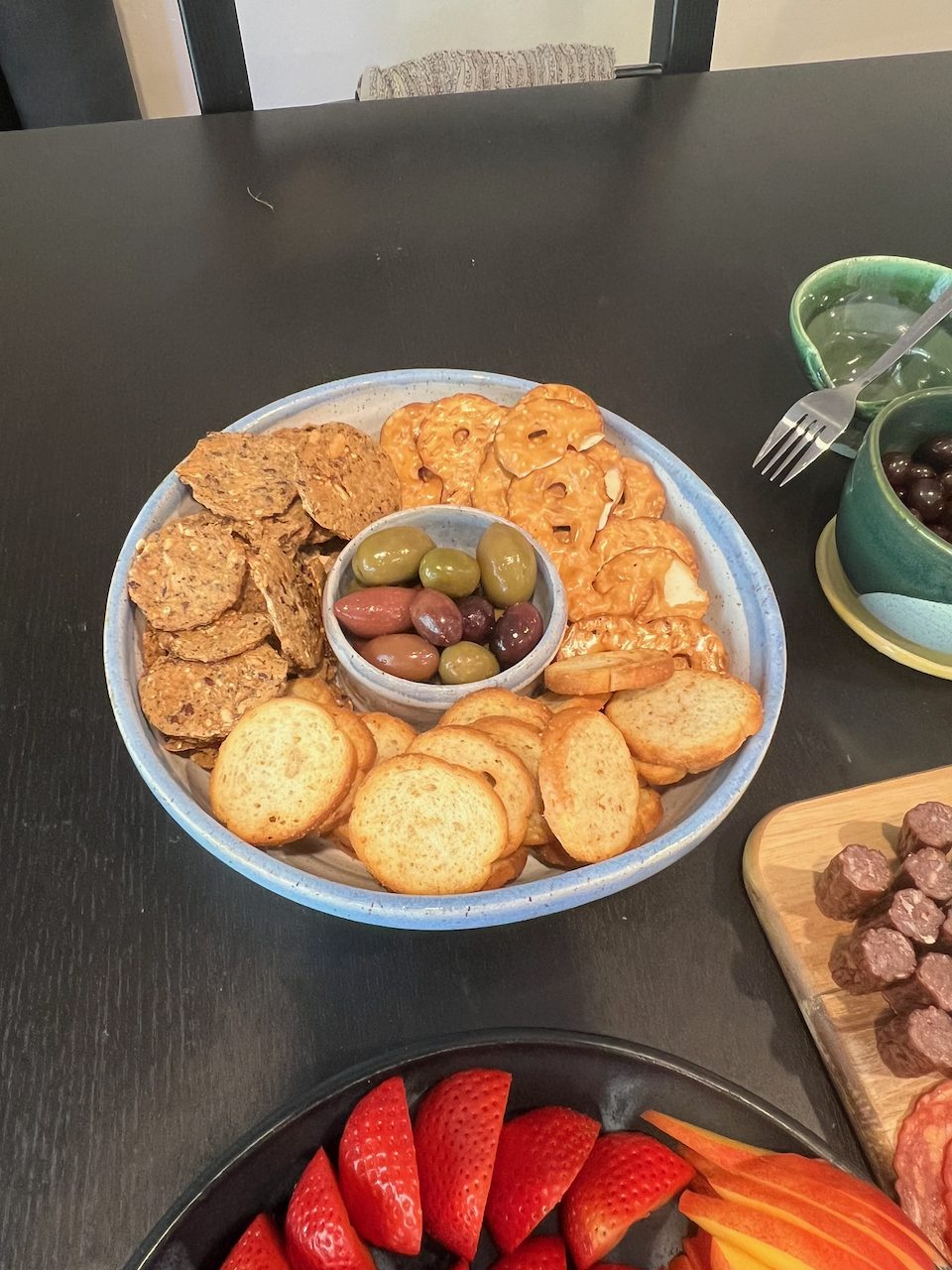 A chip and dip bowl with crackers, pretzels, and mixed olives.