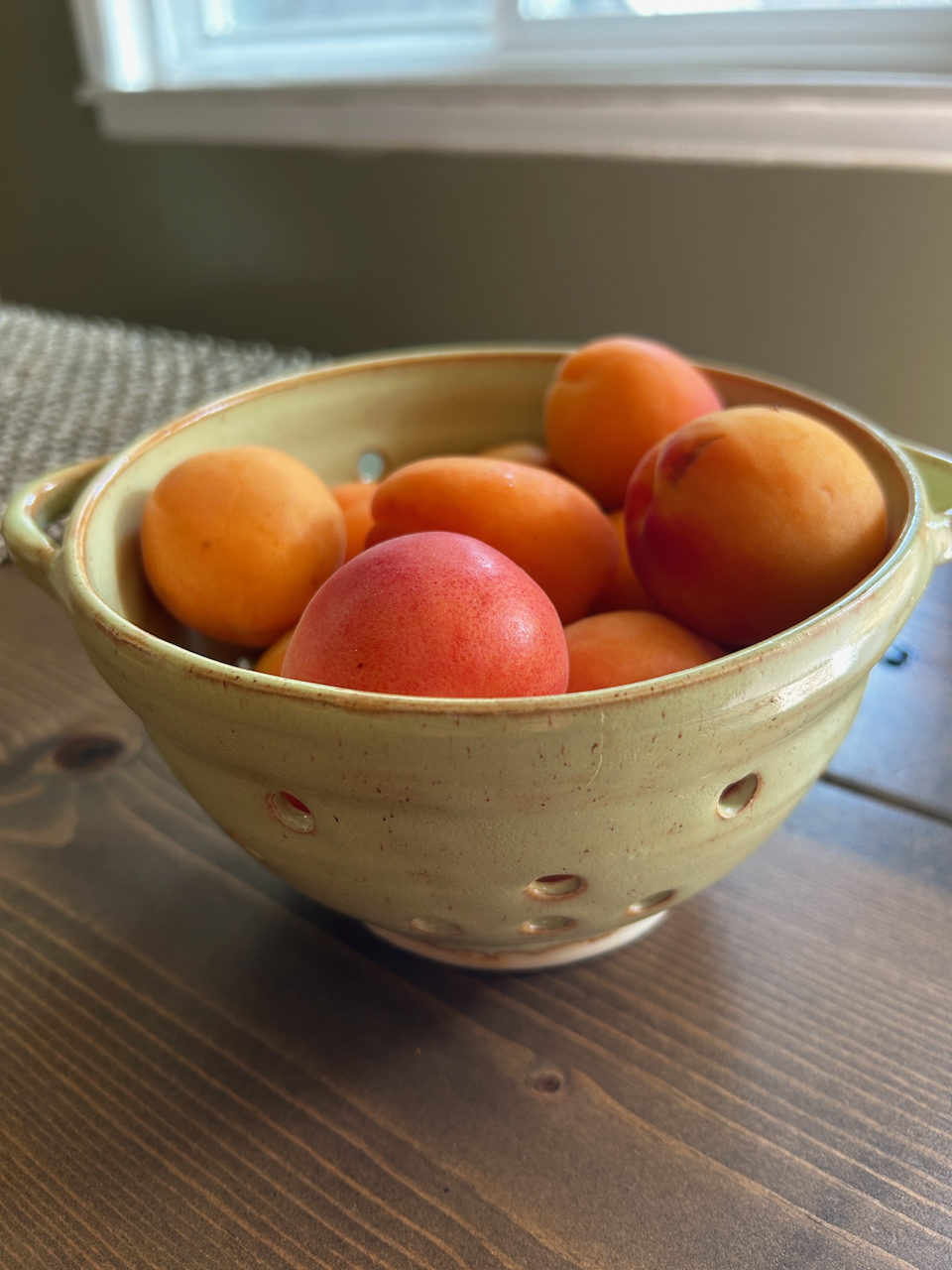 A ceramic colander filled with fresh peaches sitting on a wooden table near a window.