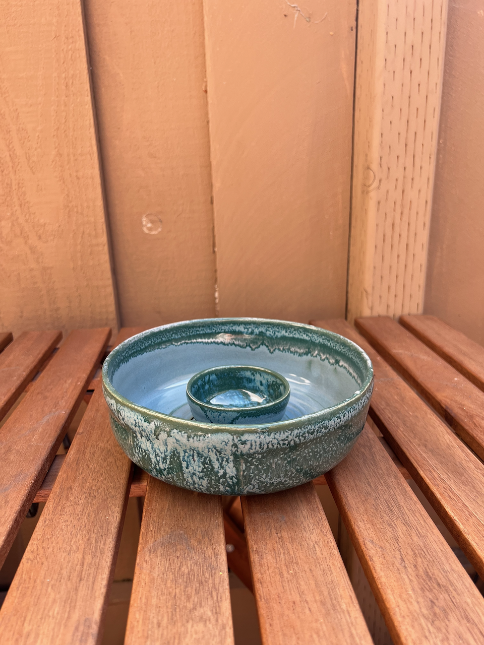 A ceramic chip and dip bowl glazed in shades of green and blue, sitting on wooden slats against a light wooden wall.