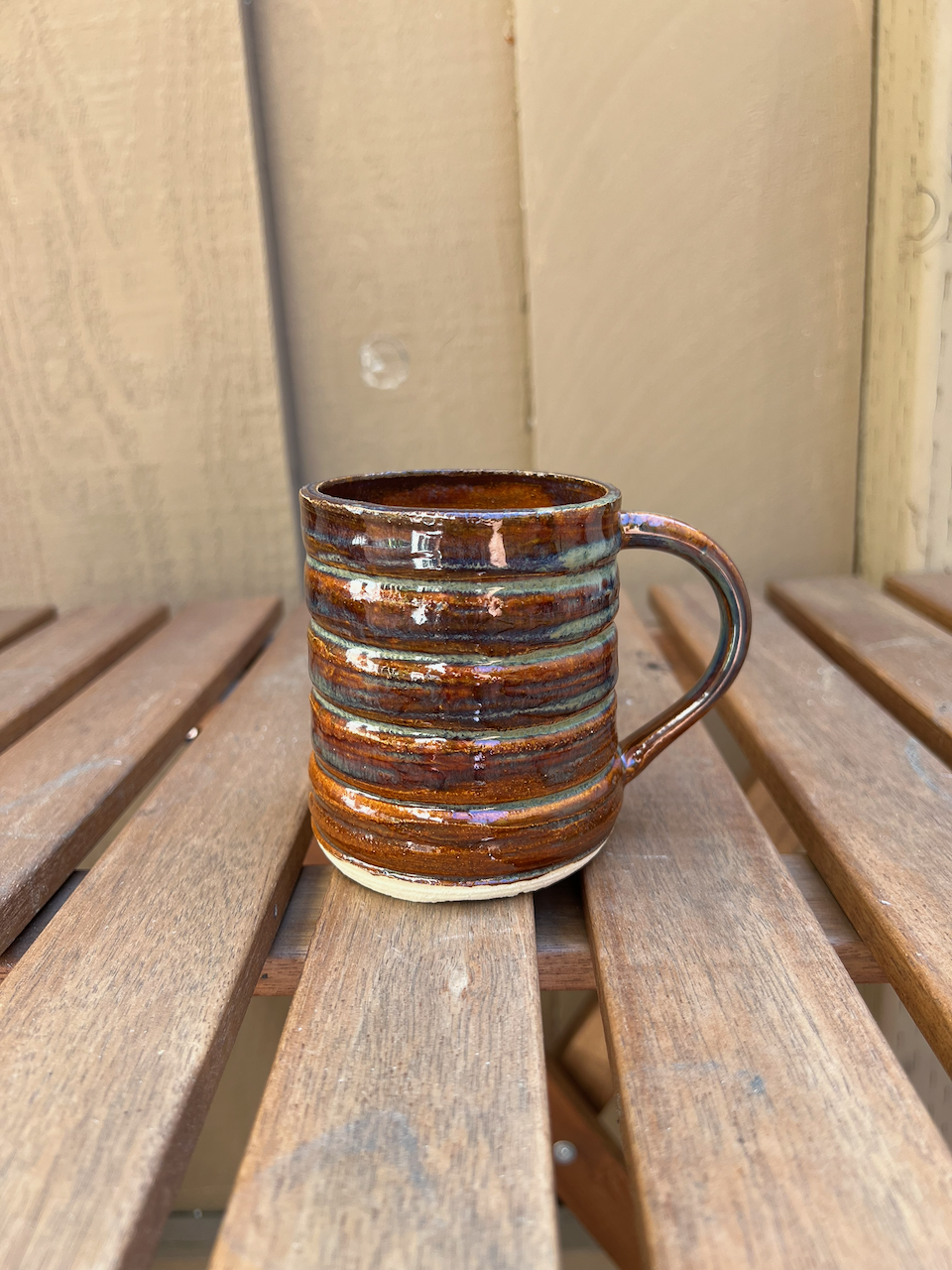 A ceramic mug with a textured, horizontal striped design, glazed in brown and blue tones, sitting on a wooden slatted surface against a beige wooden wall.