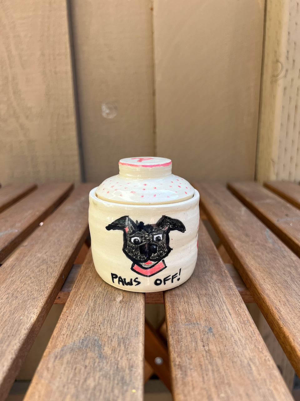 Handmade ceramic jar with a painted dog face and the words "PAWS OFF!" on a wooden surface.