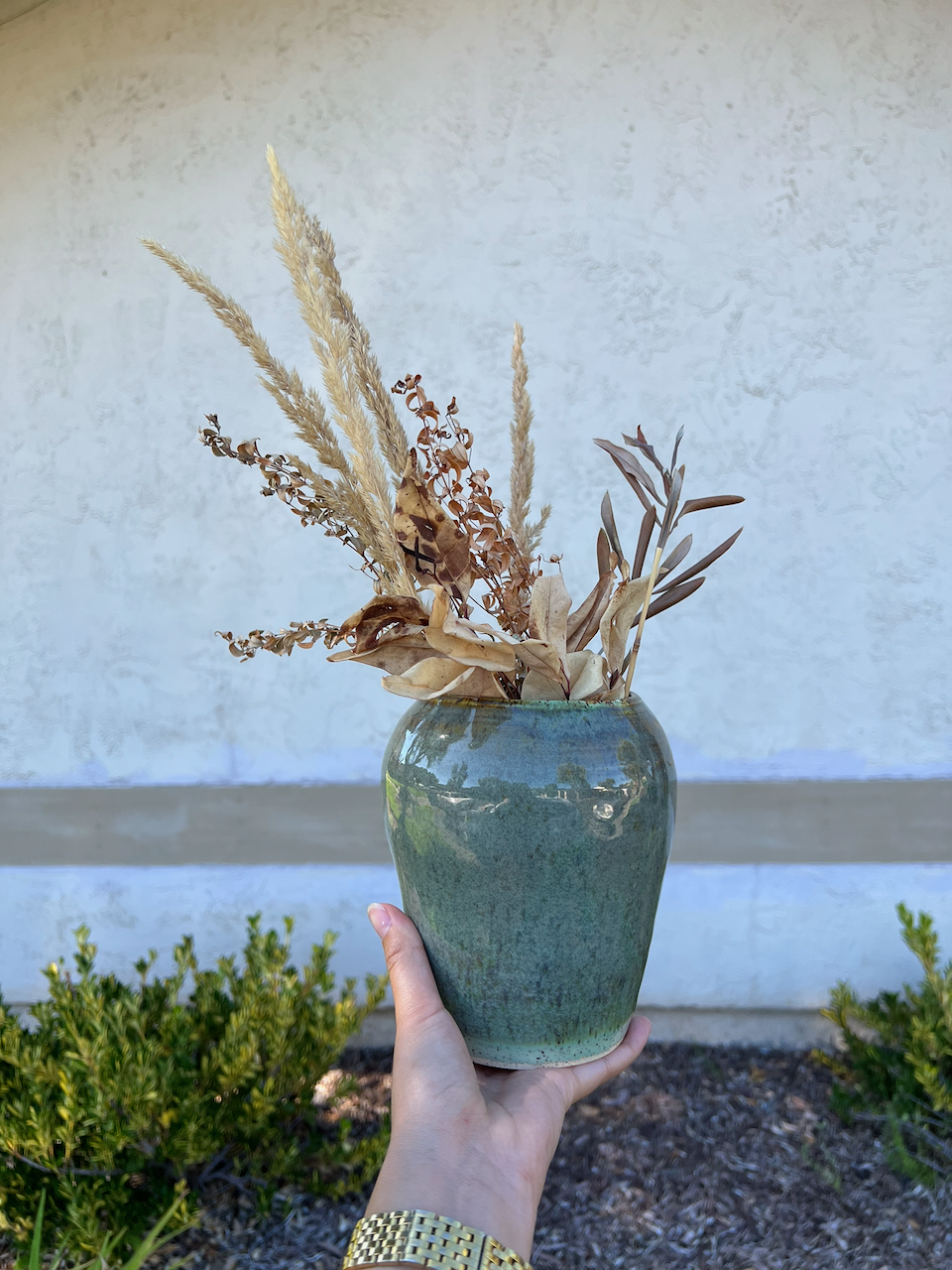A person holding a ceramic vase with dried decorative plants, including pampas grass and leaves, against a plain wall and shrubbery background.