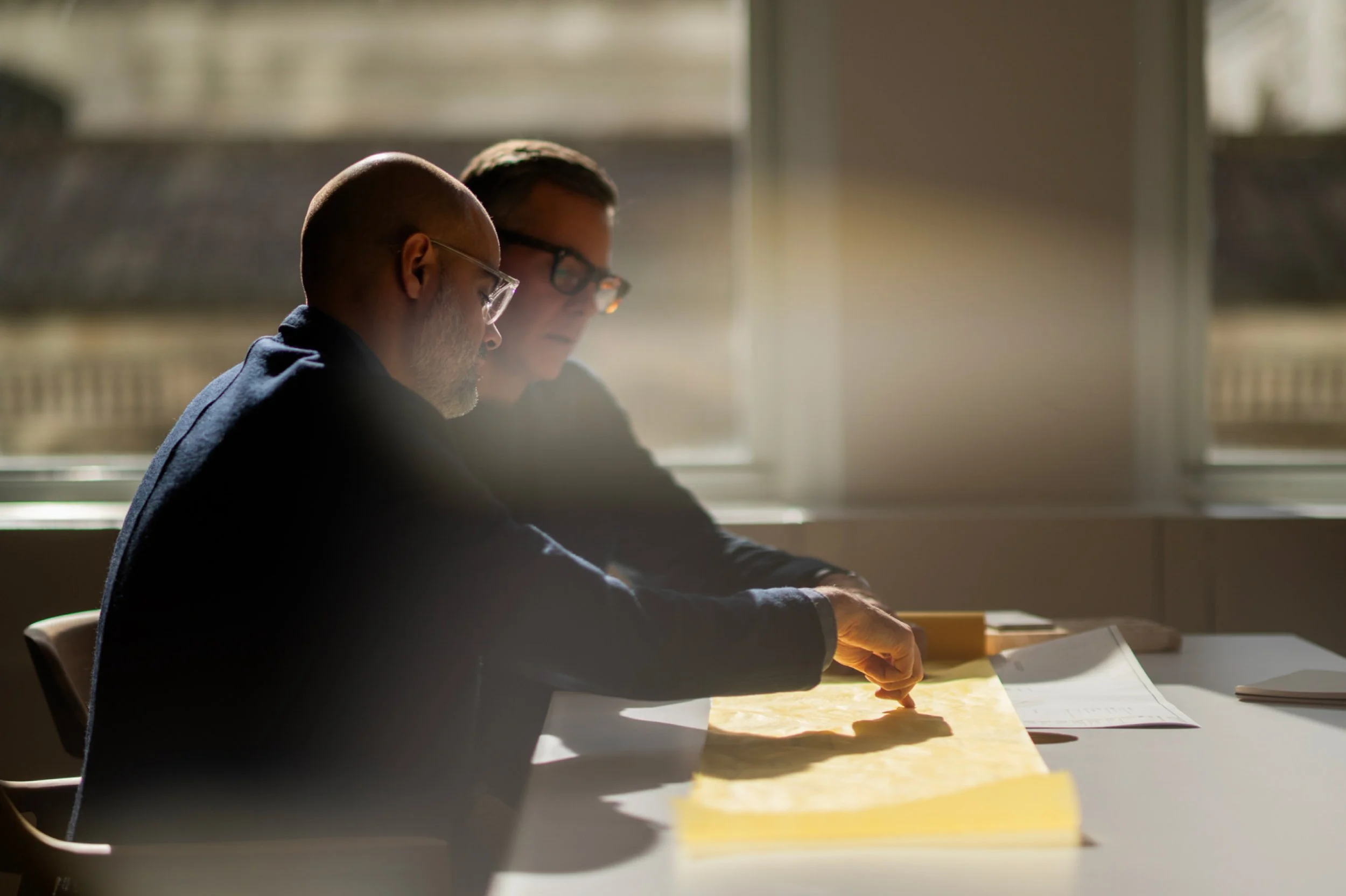 Two men sitting at a table looking at a large yellow map with documents on the side, near a window with sunlight streaming in.