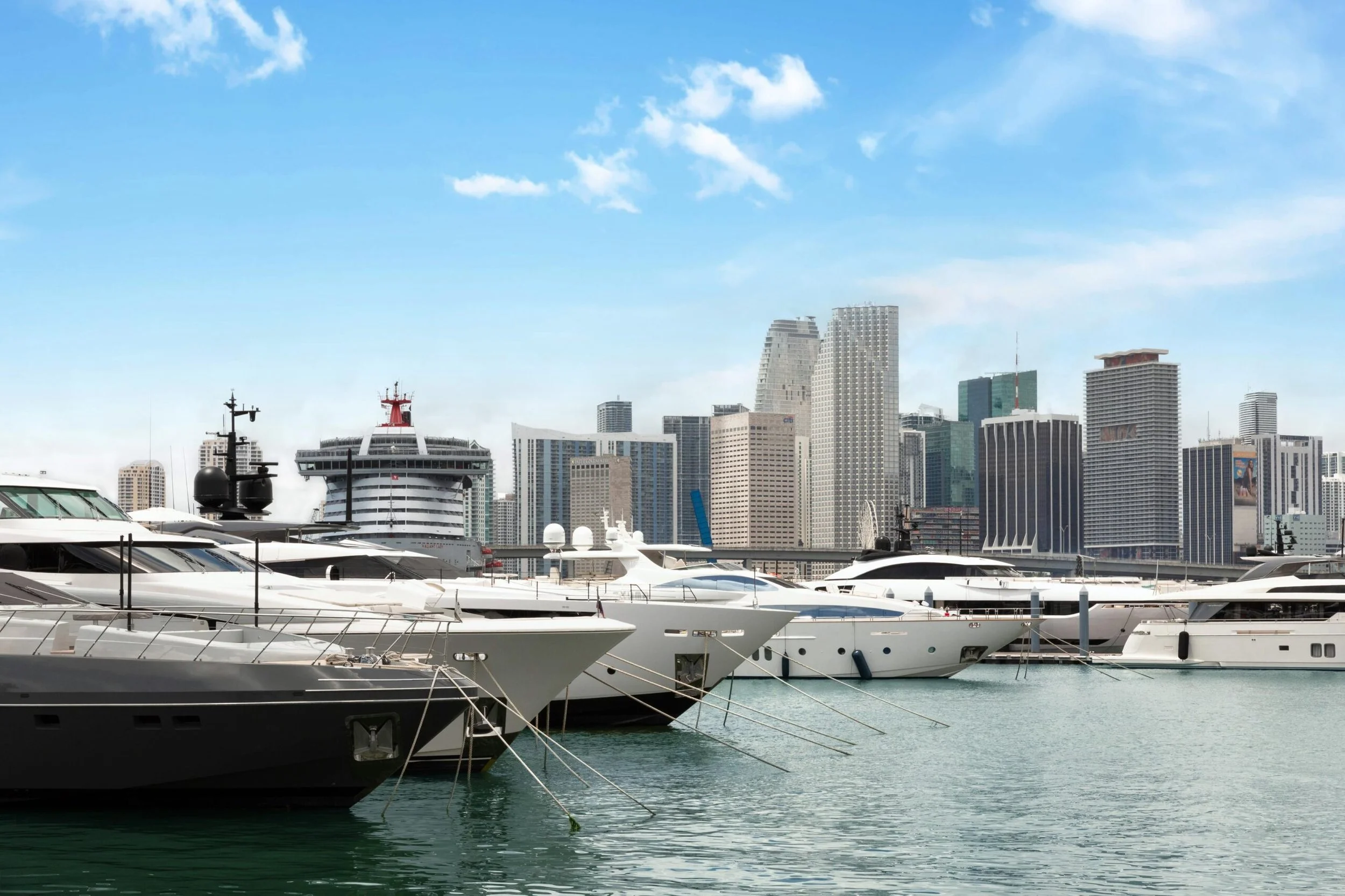 Marina with luxury yachts docked, city skyline with tall buildings in the background, clear blue sky.