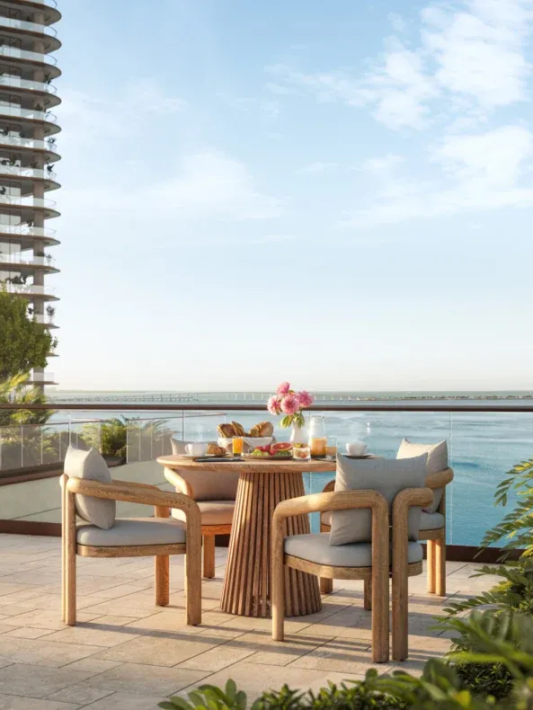 A balcony with a round wooden table set for breakfast, with four cushioned chairs, overlooking a body of water and a city skyline in the background.