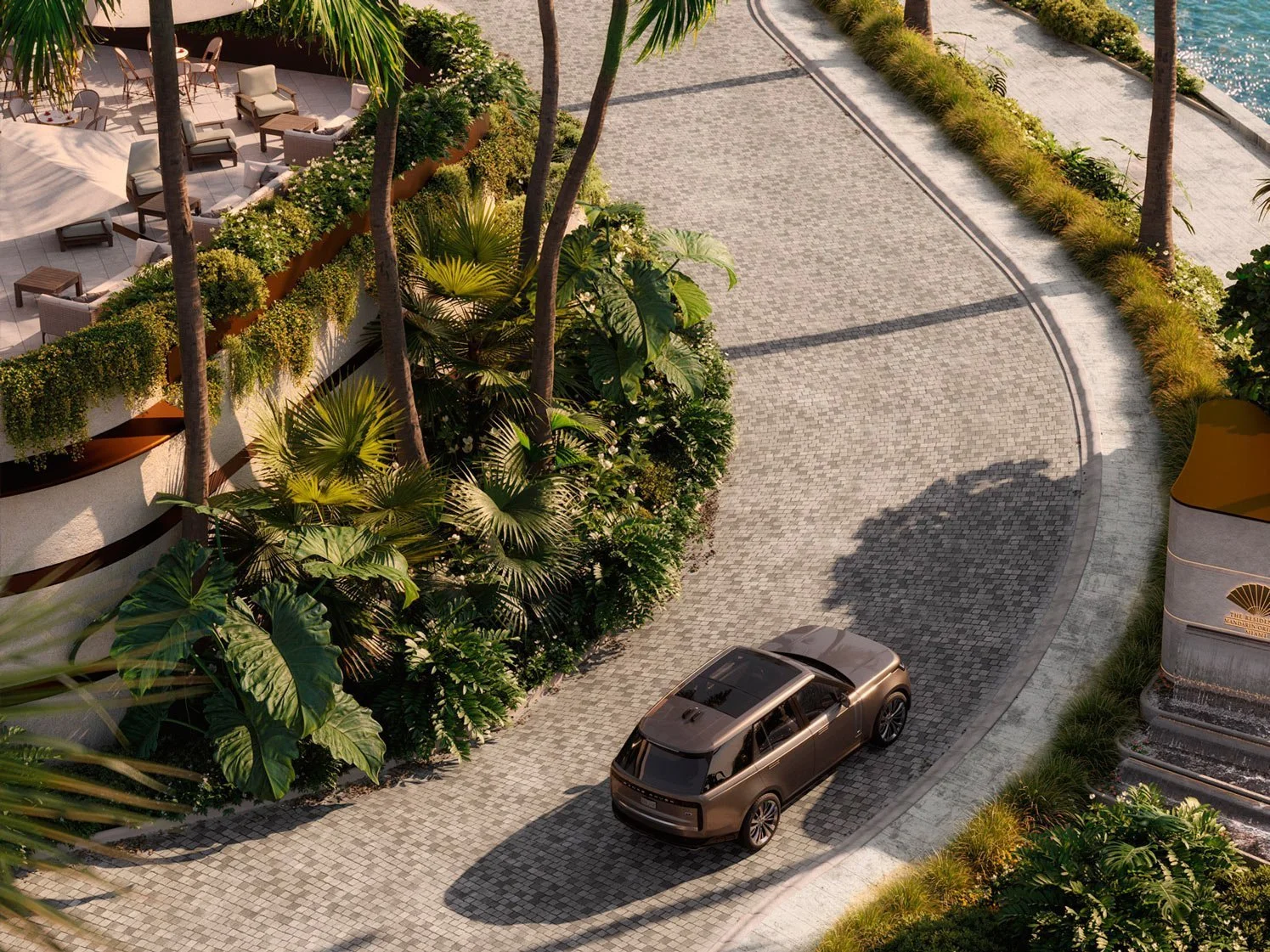 An aerial view of a luxury vehicle driving along a curved cobblestone driveway in a tropical setting. There are lush green plants and palm trees lining the driveway, and a landscaped area with outdoor seating and umbrellas nearby.