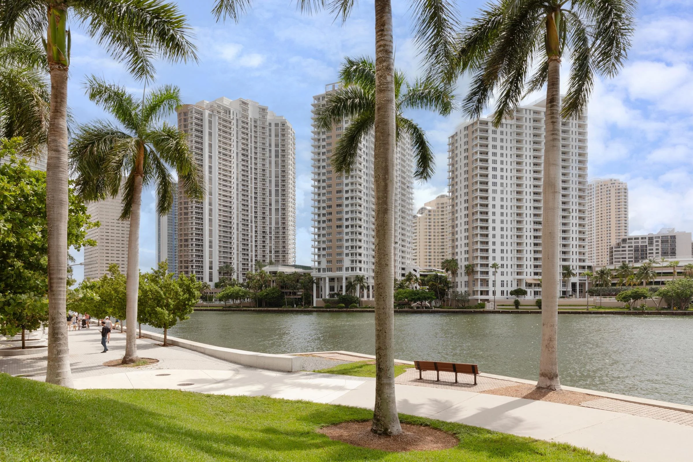 View of high-rise apartment buildings along a river with palm trees and a grassy park area in the foreground.
