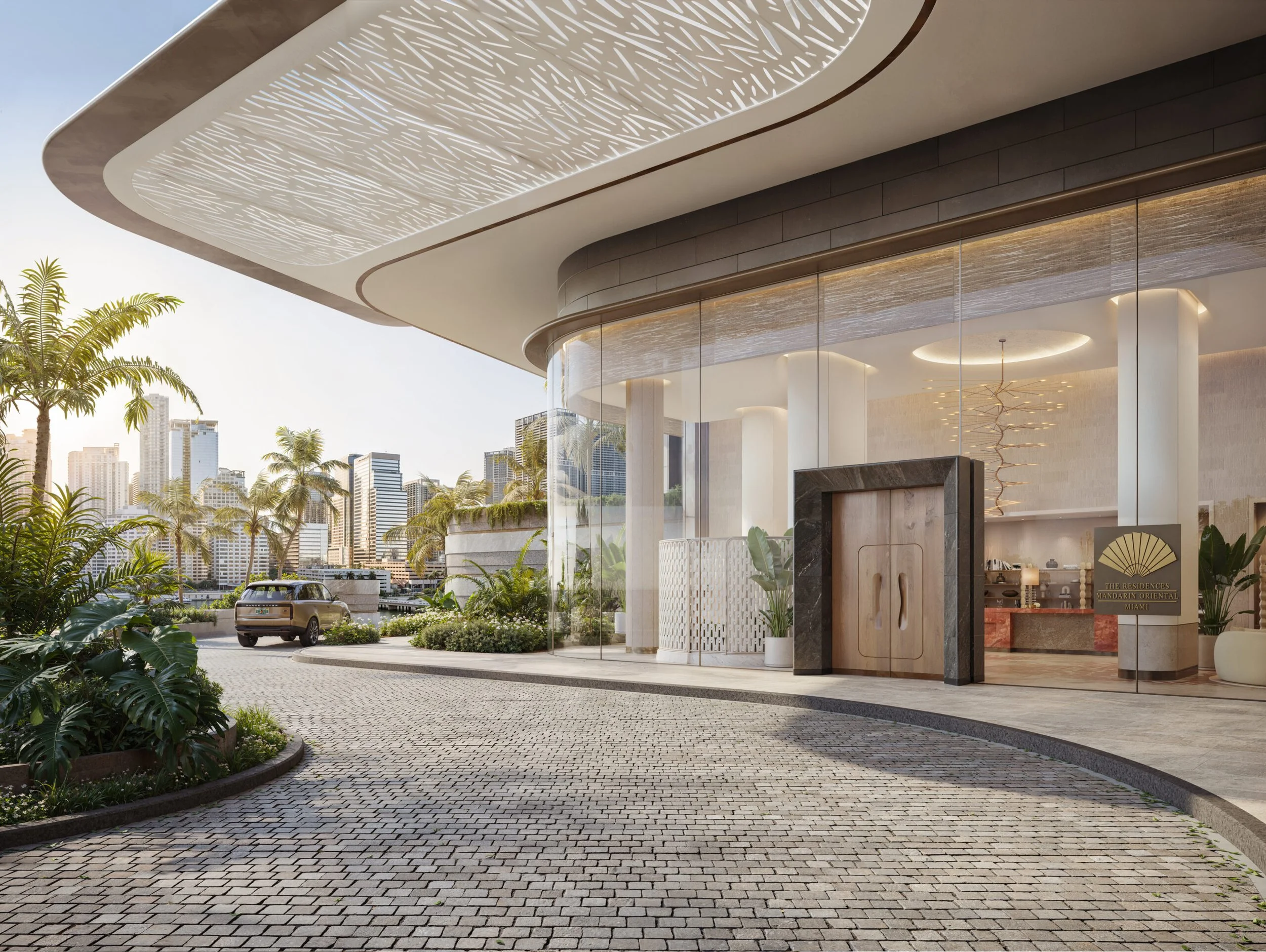 Modern building entrance with a curved canopy, glass walls, and a paved driveway surrounded by lush tropical plants and palm trees, with a city skyline in the background.