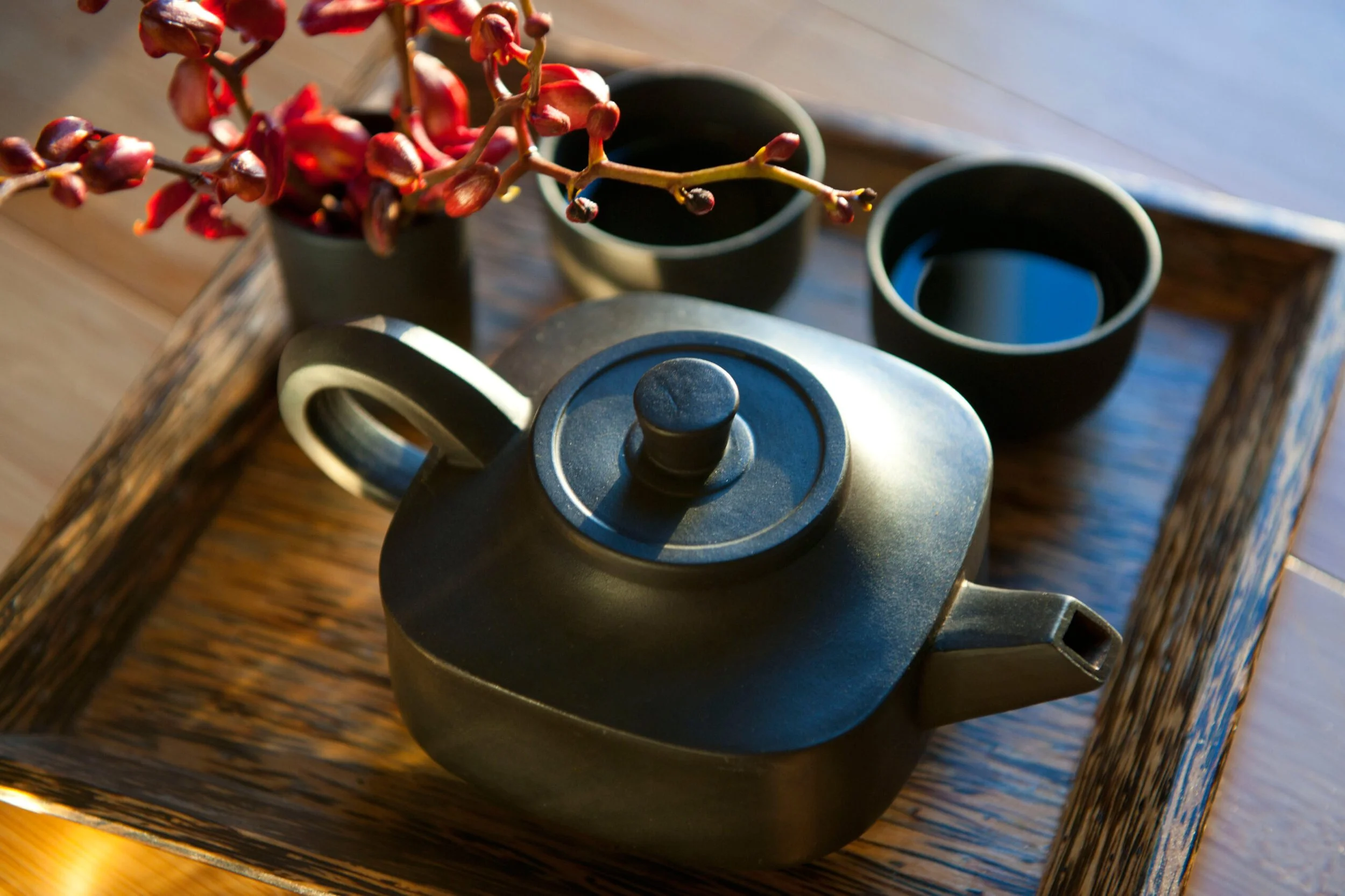 Black teapot with two black cups filled with dark liquid, sitting on a wooden tray along with a small plant with red flowers.