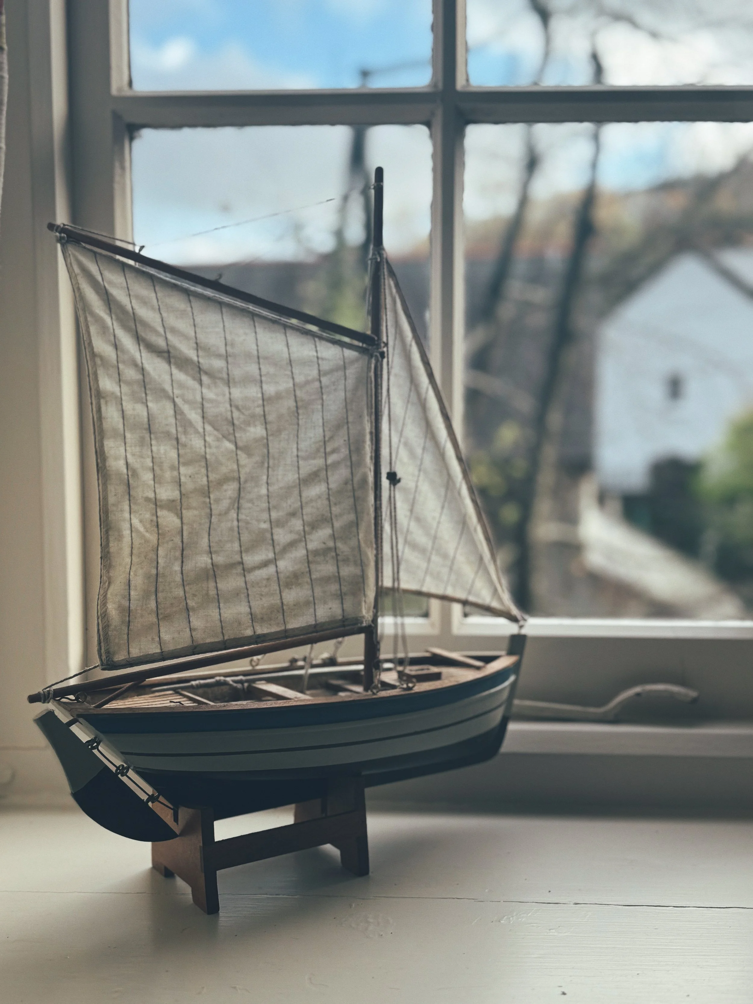 A model sailboat with a wooden hull and fabric sails is displayed indoors near a window, with a blurred background of trees and a house outside.