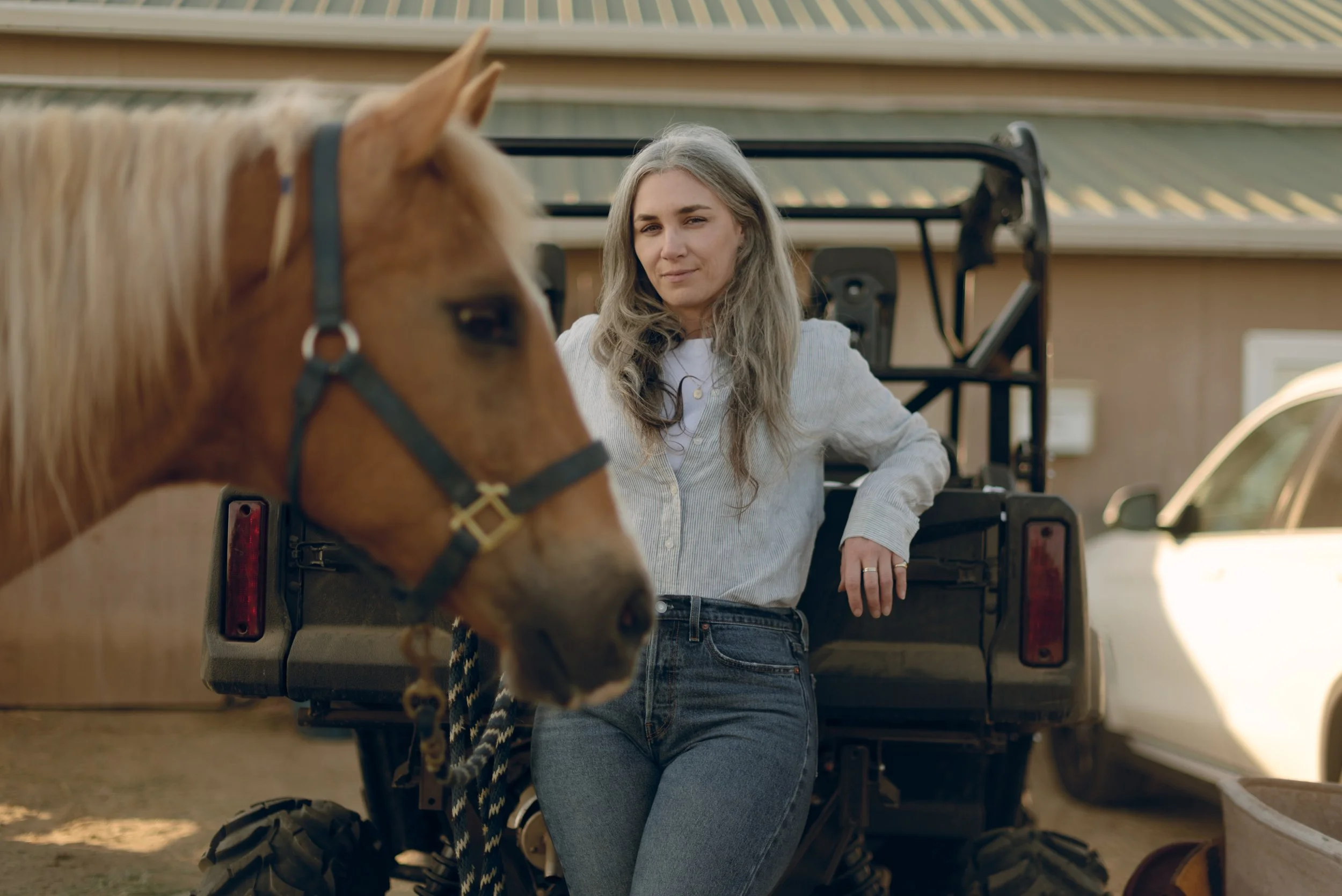 A woman with long gray hair standing next to a red horse on a trailer in a driveway.