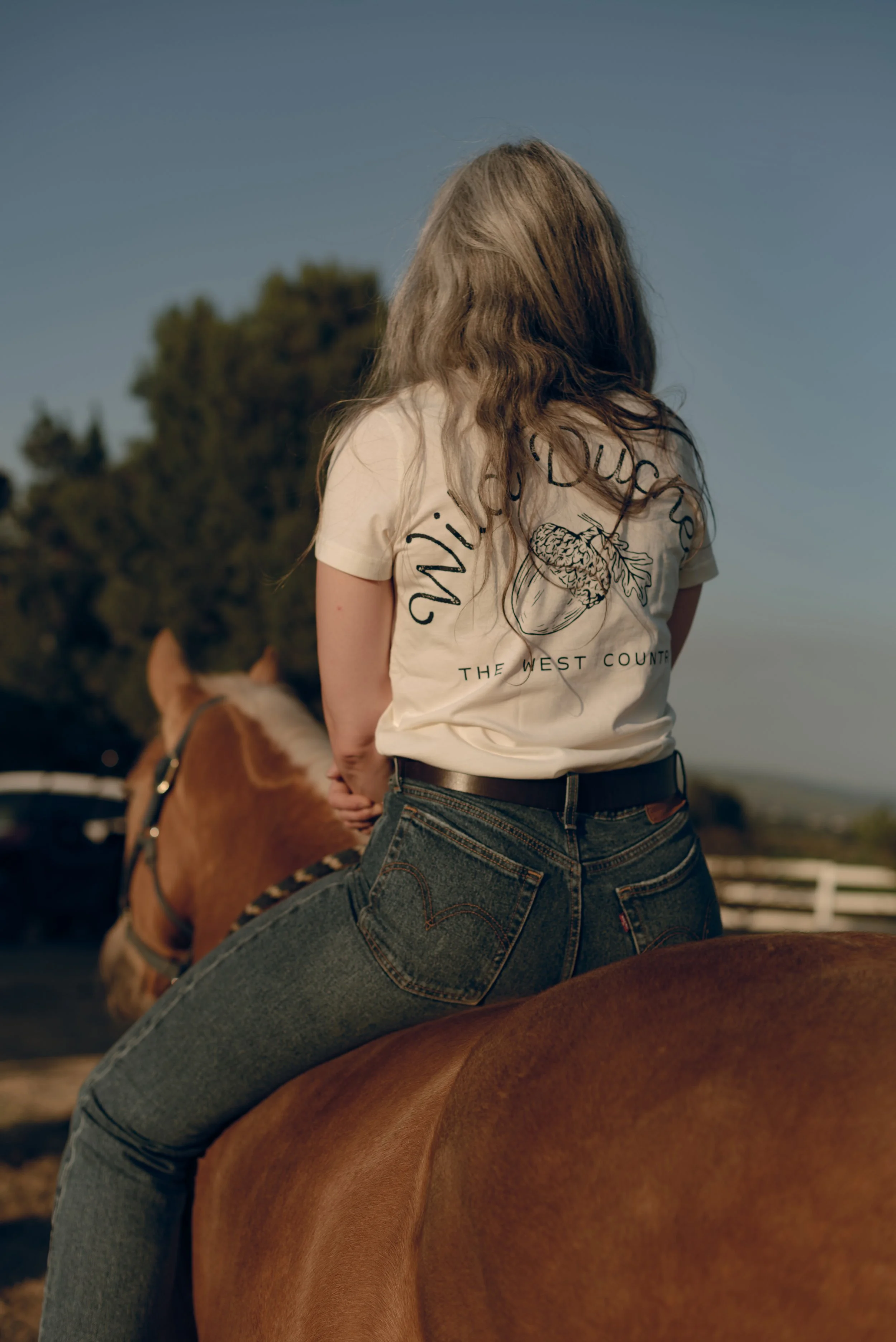 A person with wavy hair riding a brown horse during daytime, wearing a white T-shirt with a graphic and text, and jeans. The background features trees and a clear sky.