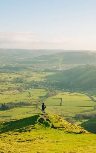A person standing on a hilltop overlooking a lush green valley with patchwork fields, hills, and a partly cloudy sky in the background.