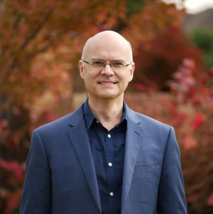 A man wearing glasses and a blue suit jacket, smiling outdoors with autumn-colored trees in the background.